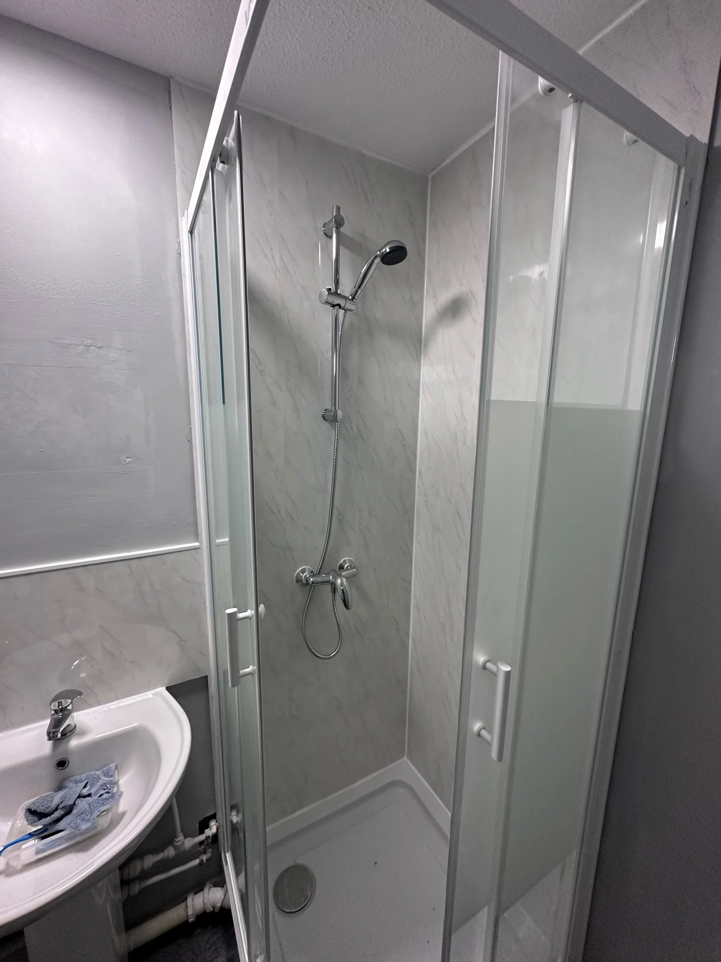 Small bathroom with a corner shower stall, clear glass door, showerhead, and marble-patterned walls, with a small sink and towel on the counter.