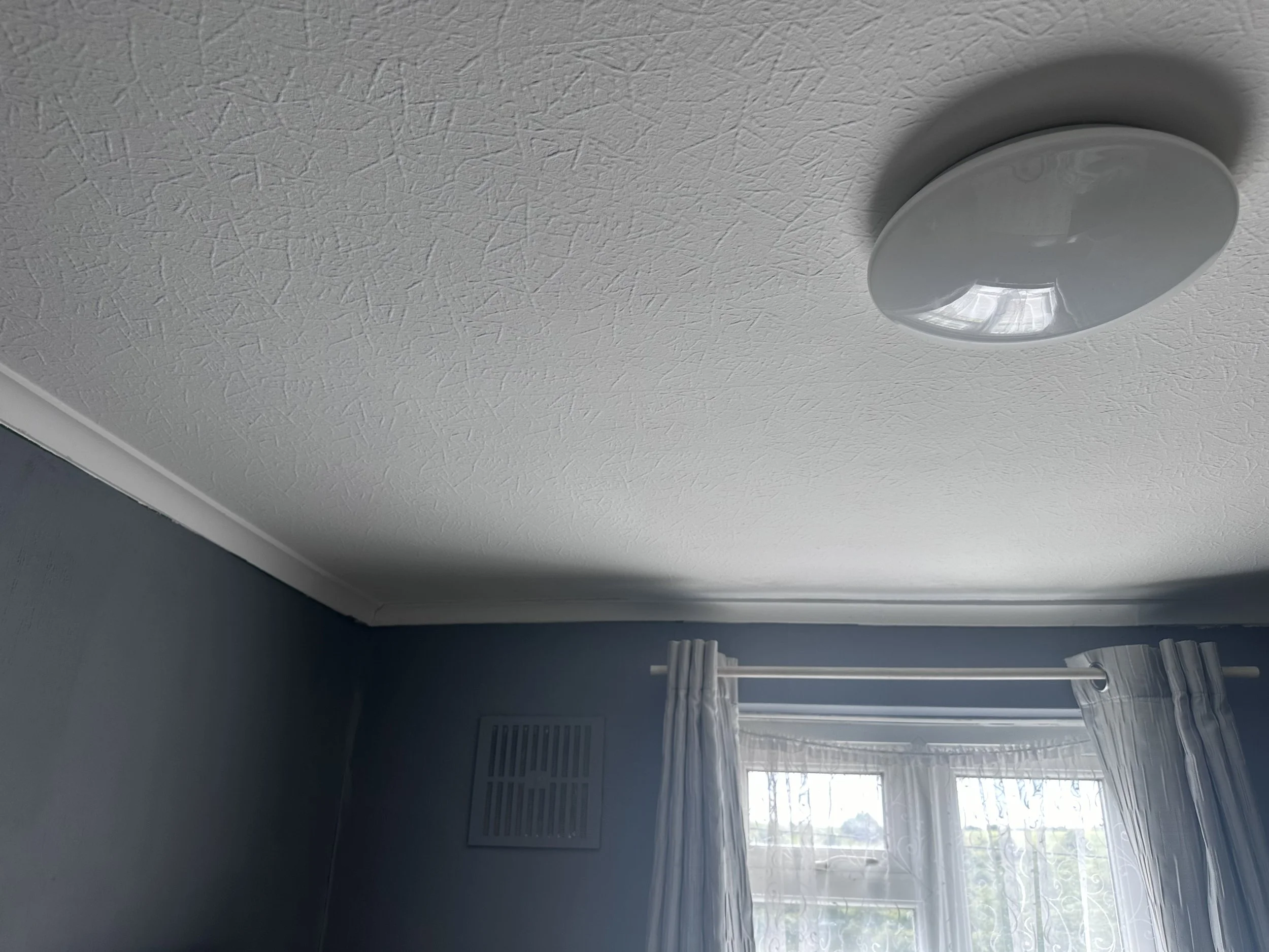 A white ceiling with a textured finish, a round white ceiling vent, and a part of a window with sheer white curtains and a window blind underneath.