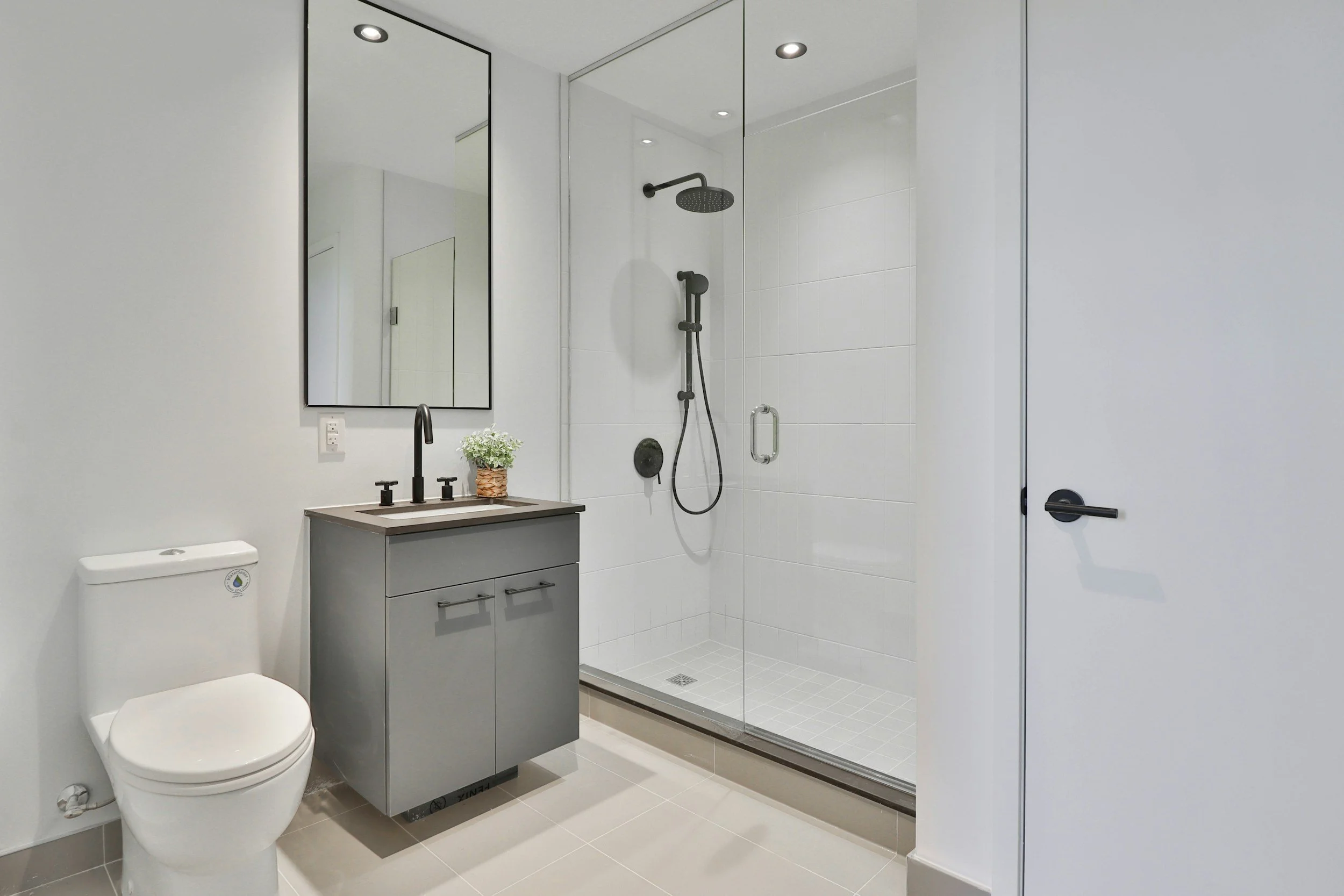 Modern bathroom with a toilet, gray vanity with black faucet, large mirror, and a glass-enclosed shower with black fixtures.