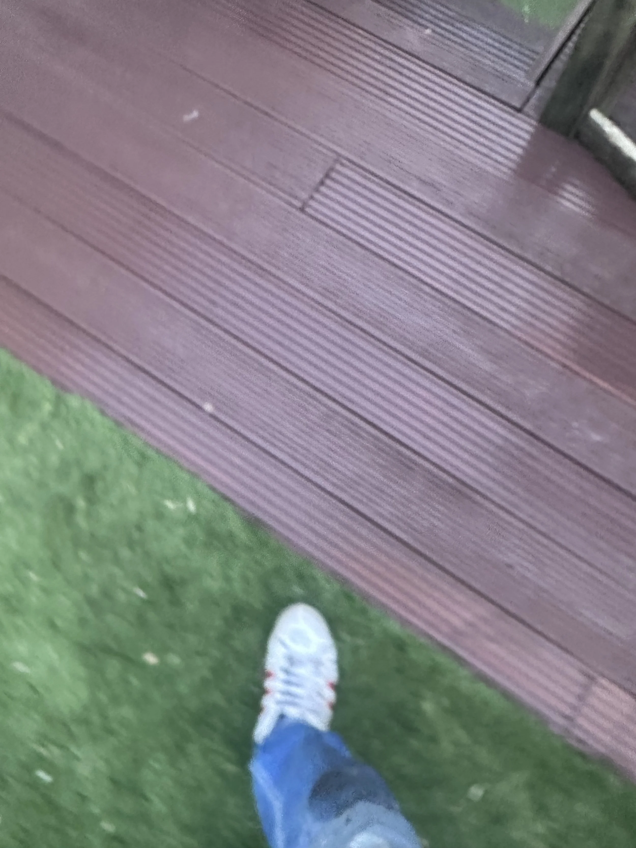 A person wearing white shoes, blue jeans, and a gray jacket is walking on a grassy area next to a wooden deck. The photo is taken from their perspective.