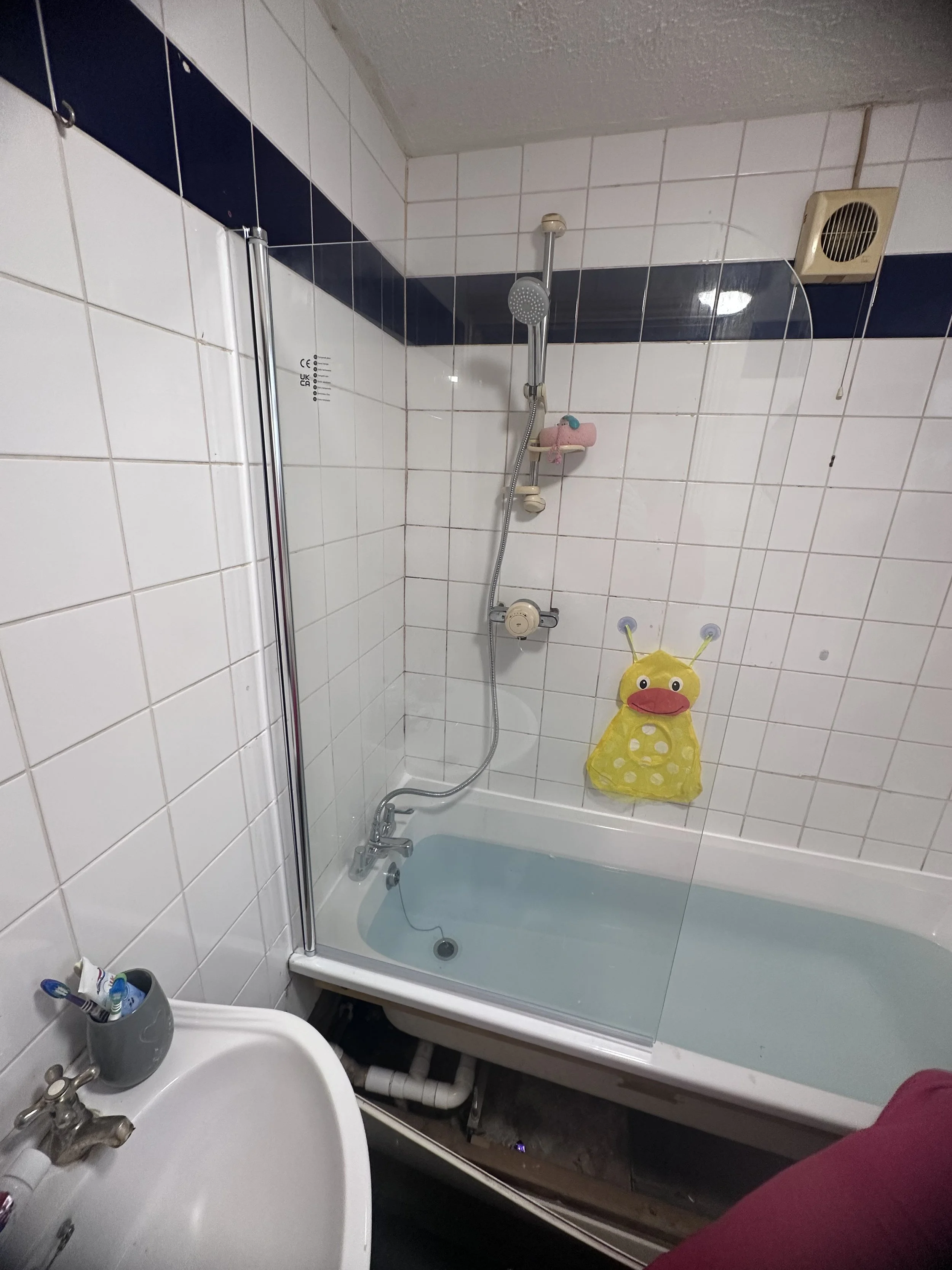 A small bathroom with a bathtub, a showerhead, and white tiled walls with a blue stripe. There is a yellow duck-shaped bath sponge hanging on the wall and a toilet with toothbrushes and toothpaste on the sink.
