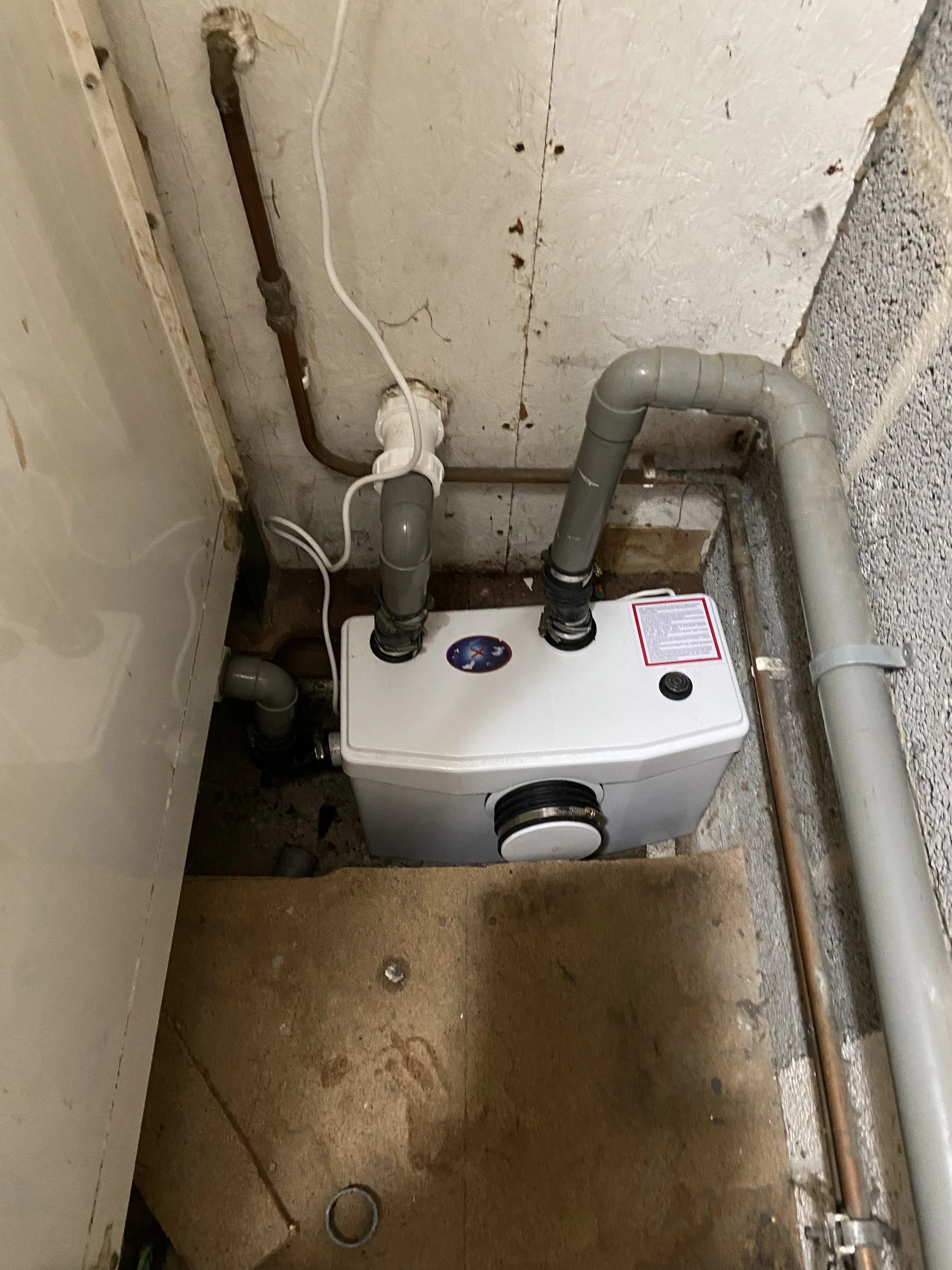The image shows a utility room with exposed plumbing and a tankless water heater installed on the wall, with a concrete floor and unfinished walls.