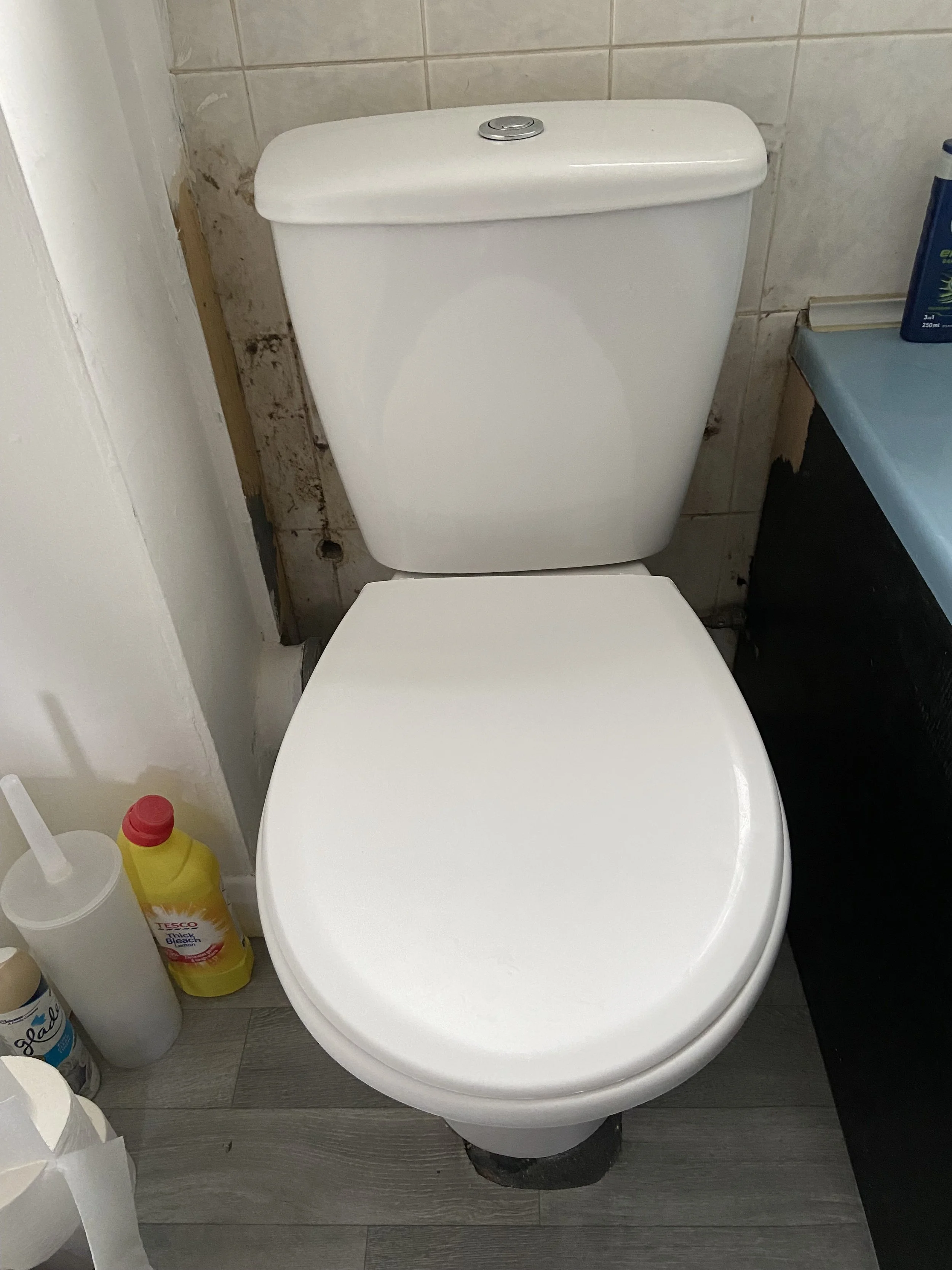 A white toilet with a closed lid, situated in a bathroom with tiled wall behind it and wooden flooring below. Several cleaning supplies are visible to the left and right of the toilet.
