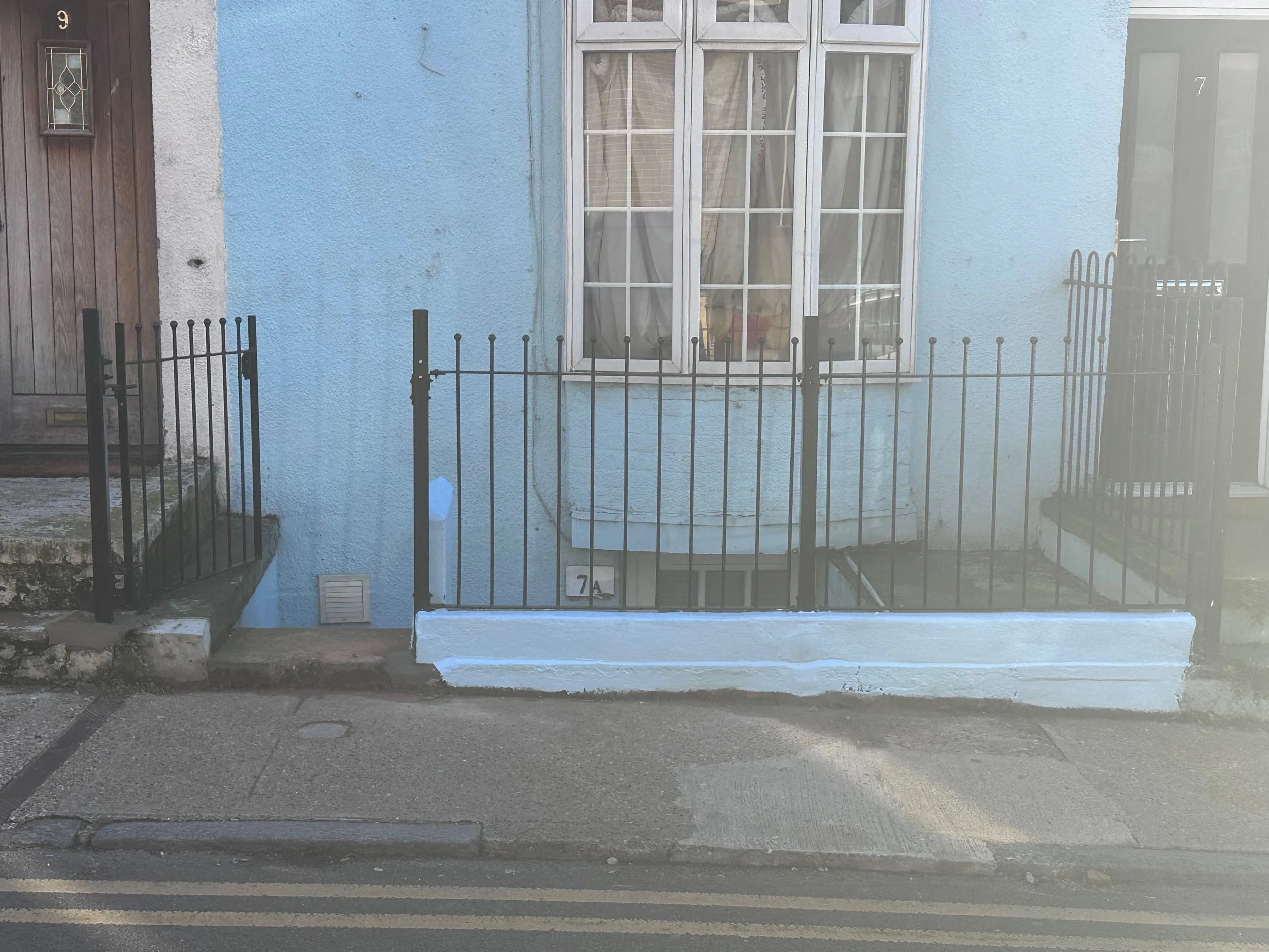 Front of a house with a light blue exterior wall, a large window with divided panes and curtains, a black metal fence, a wooden front door, and house number signs 7 and 7A.