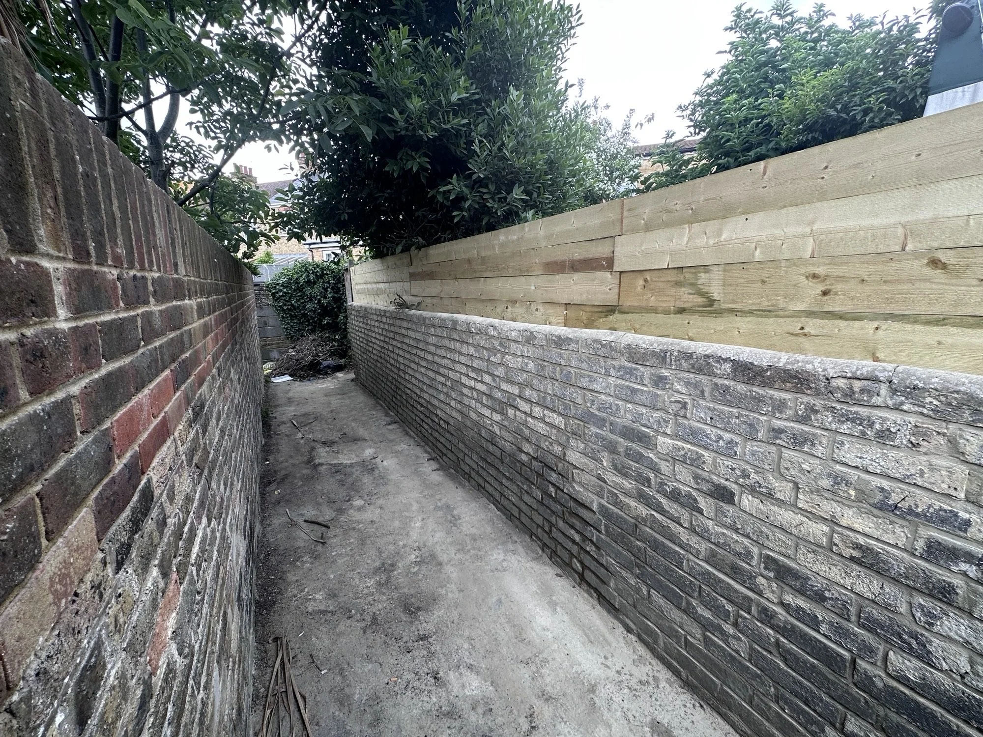 A narrow alleyway with brick walls on both sides, one side with red bricks and the other with gray bricks, and a dirt floor. There are trees and bushes at the end of the alley.