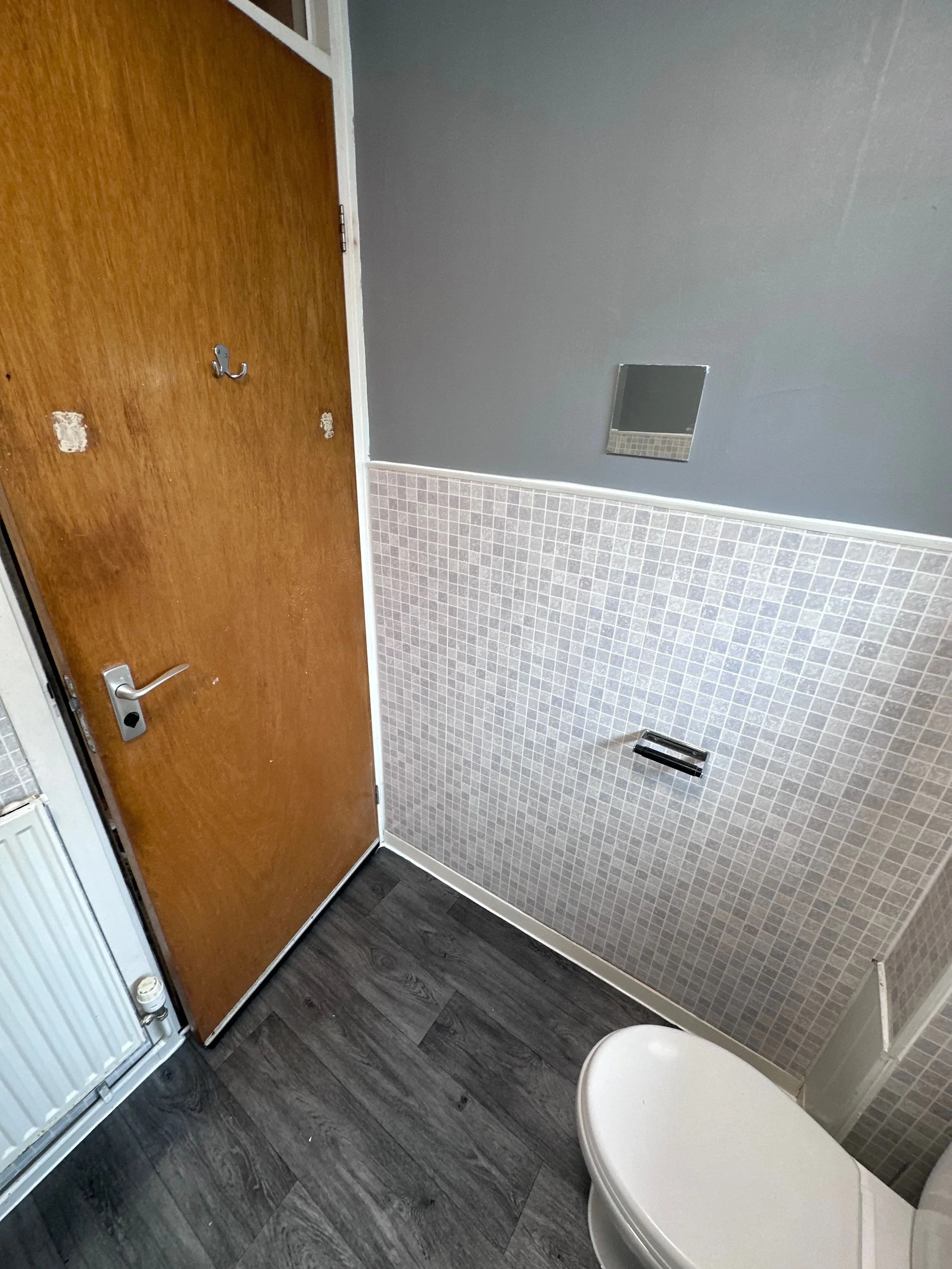 Bathroom corner with a wooden door, a small mirror on a gray wall, square beige tiles below the mirror, a bathtub at the bottom right, and dark wood flooring.