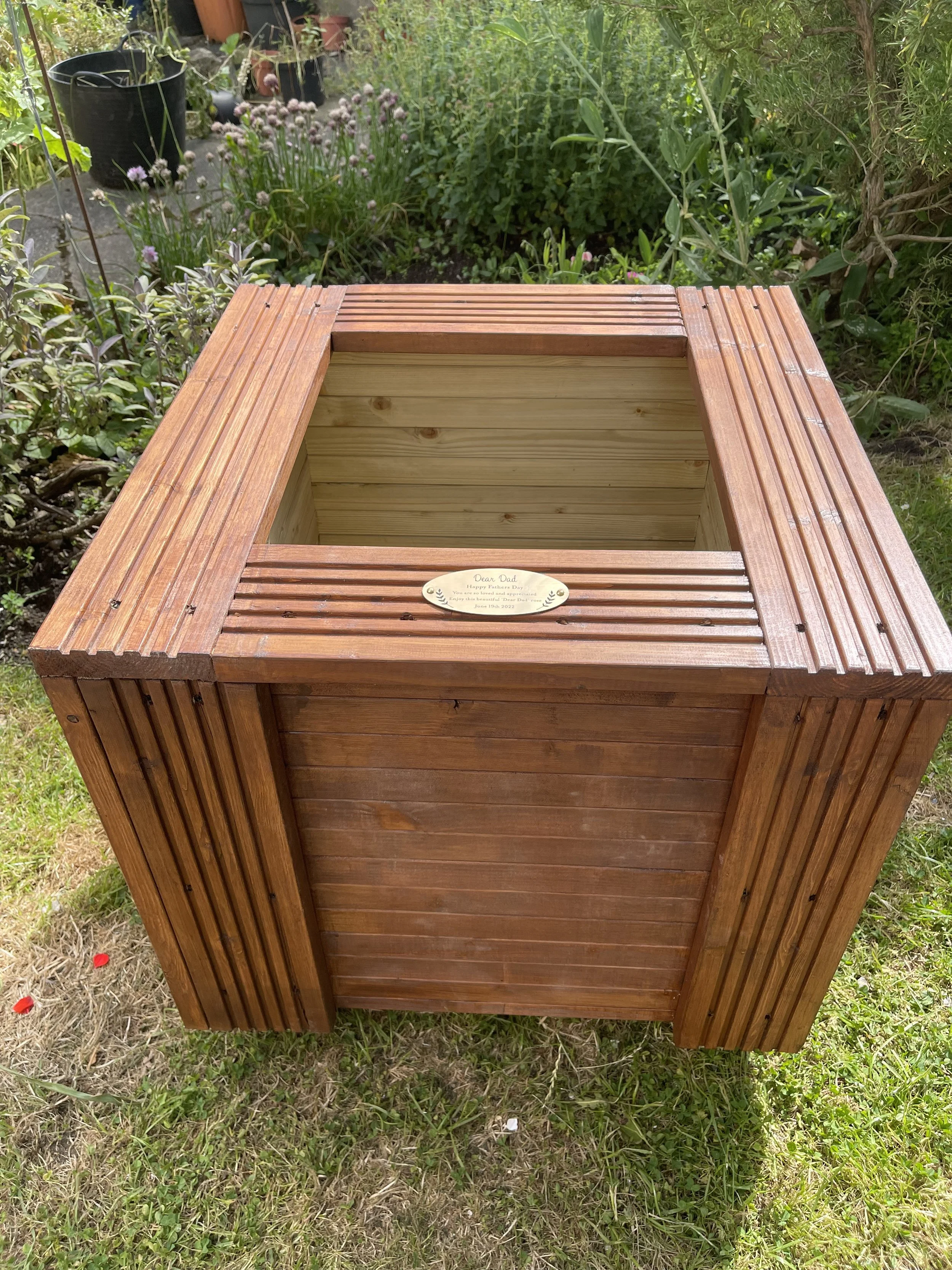 A wooden raised garden bed with a small plaque on top that reads "Dear Dad, Happy Father's Day, 2022, Enjoy this beautiful garden, June 19, 2022." The garden bed is outdoors surrounded by green foliage and potted plants.