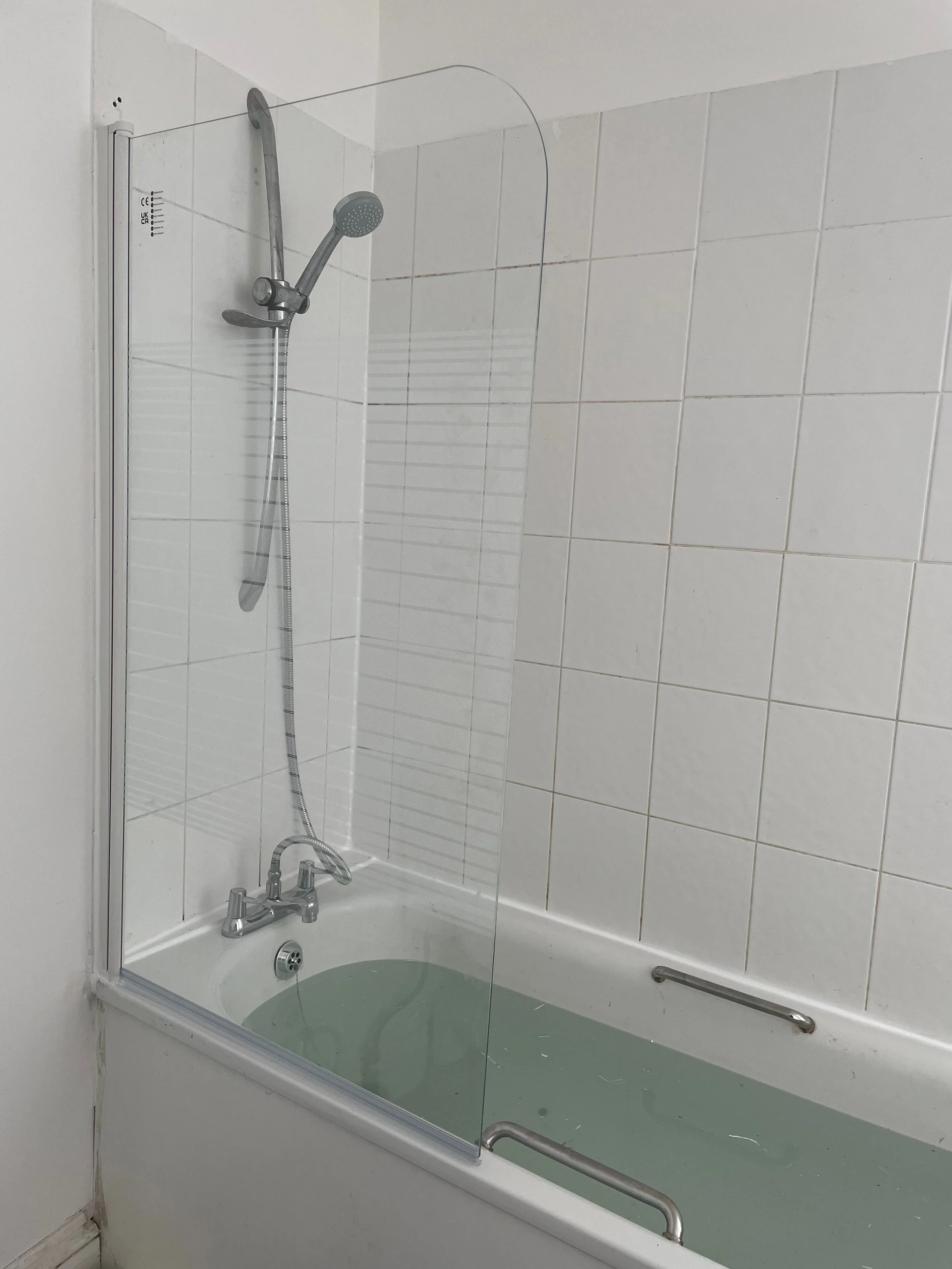 Bathroom with white tiled walls and a bathtub filled with greenish water, with a glass shower screen, showerhead, and metal grab bar.