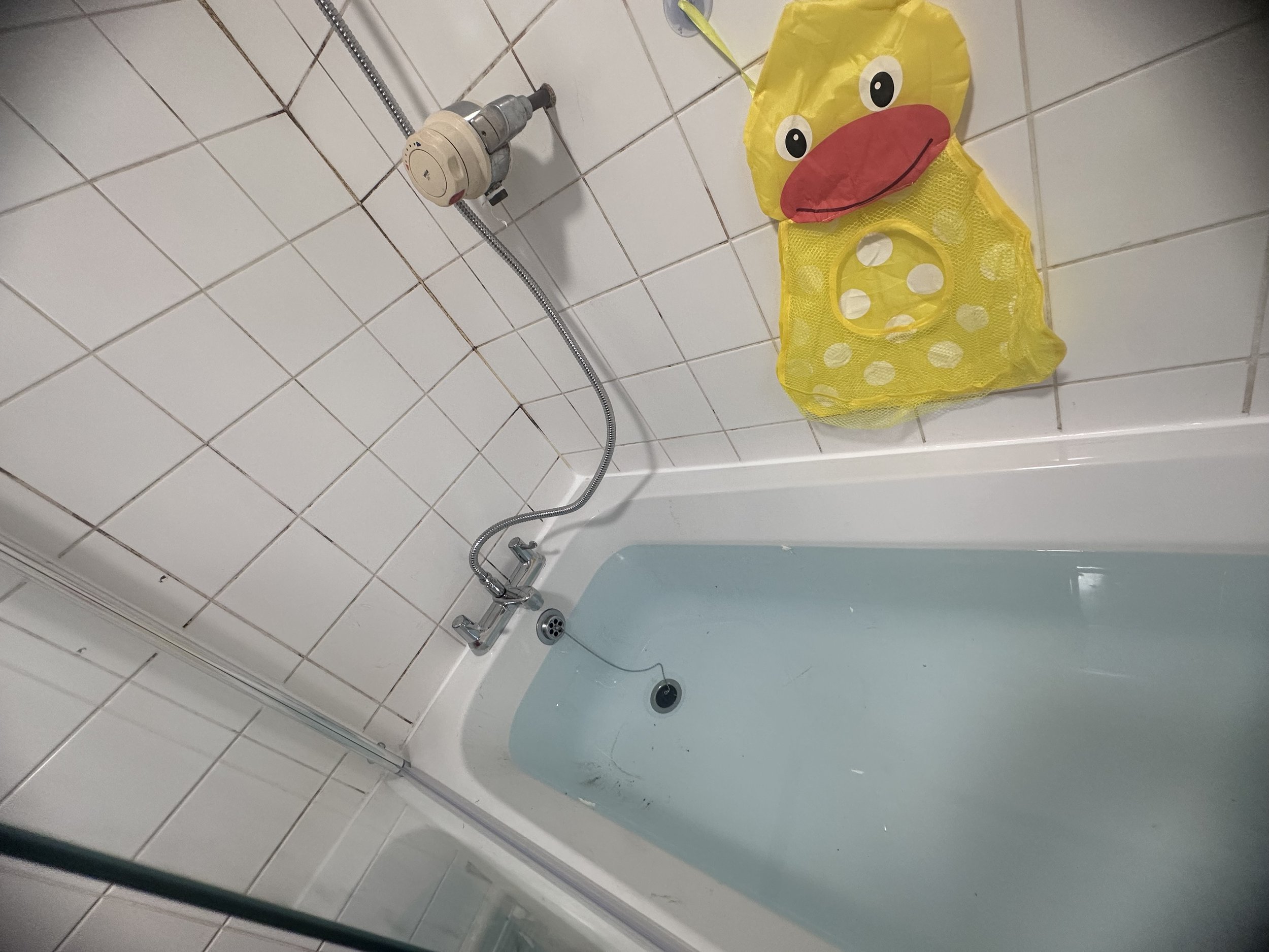 A bathroom with white tiled walls and a bathtub filled with water. A yellow duck-shaped bath toy with a red beak and big eyes is on the edge of the bathtub. There is a showerhead, a silver hose, and a control knob on the wall.