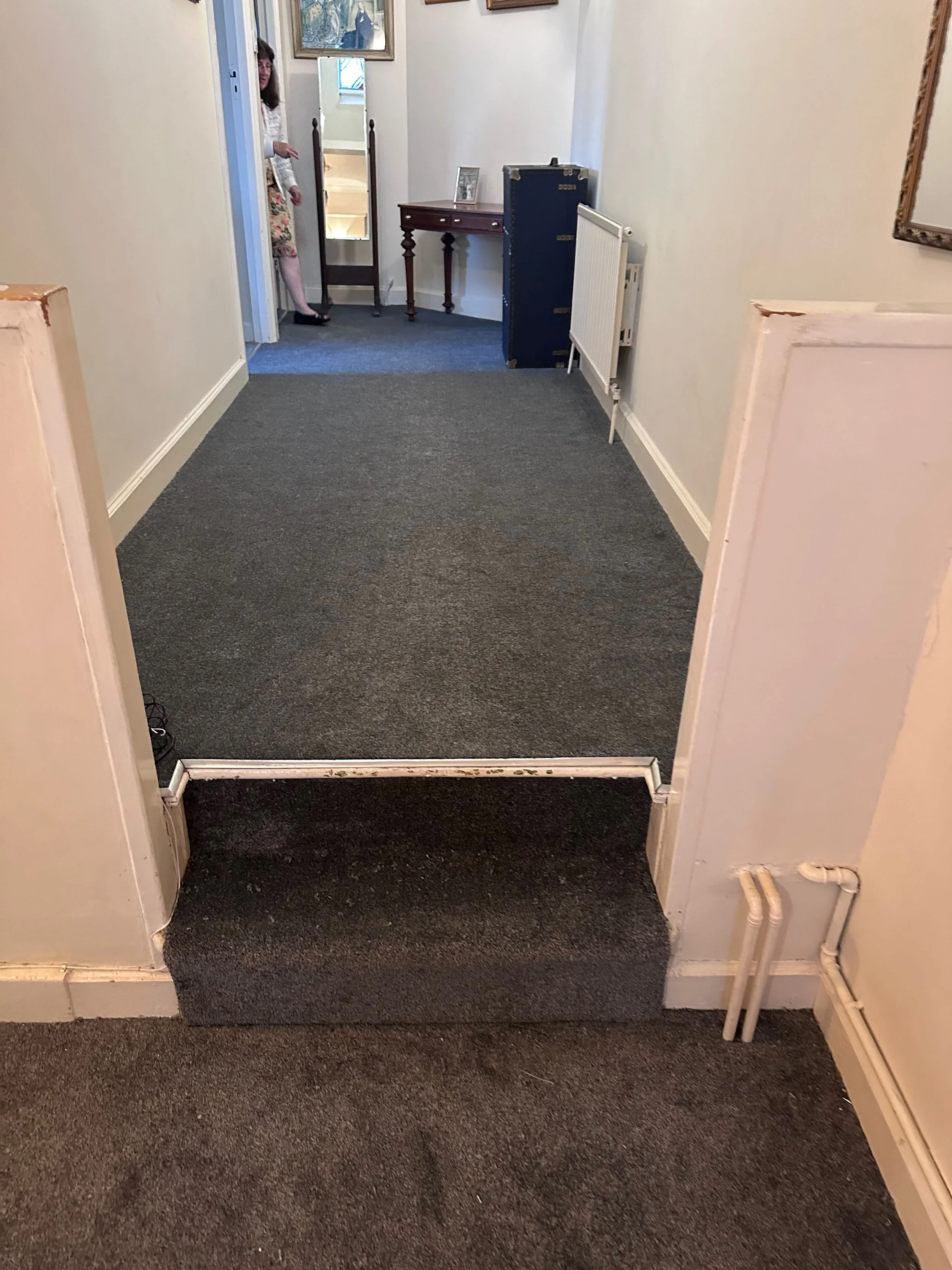 A hallway with a grey carpet and a door frame in the foreground. There is a radiator on the right wall and some exposed pipes near the bottom right corner. The hallway leads to a room with a small wooden table, a standing mirror, and framed pictures on the wall. A woman is standing in a doorway in the background, partially visible, wearing a floral skirt and a white cardigan.