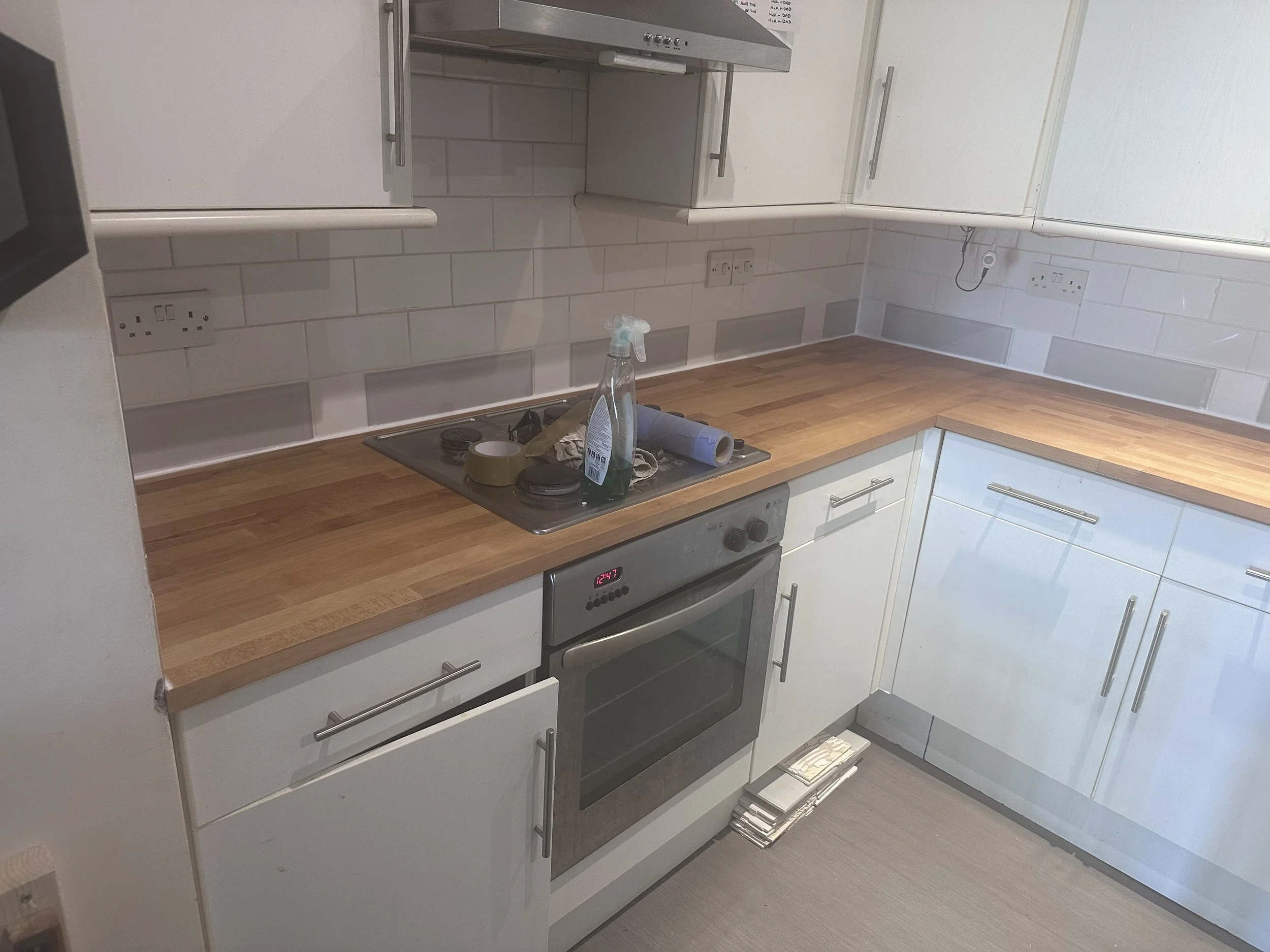 A kitchen with white cabinets, a wooden countertop, a stove with a black cooktop, and some cleaning supplies on the counter. The backsplash has white tiles, and there are multiple power outlets on the wall.