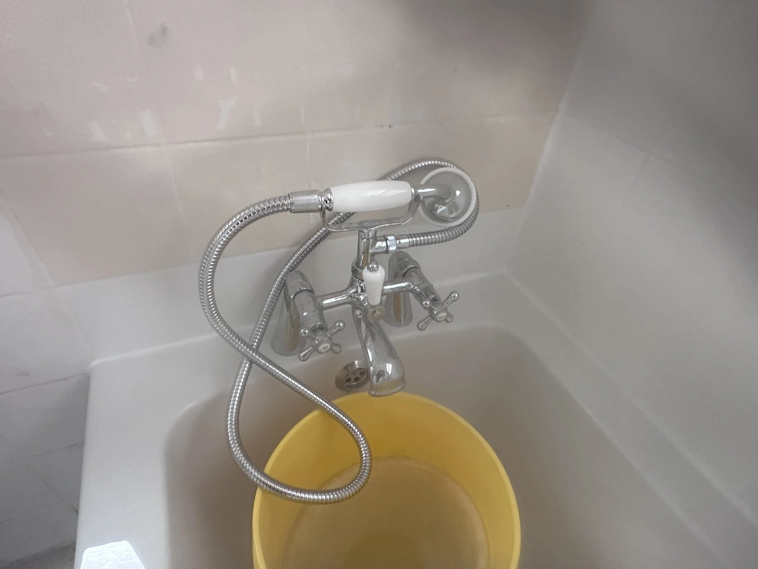 A bathroom with a white bathtub and a silver shower head attached to the wall. A yellow bucket is inside the tub and a yellow container is on the floor nearby. The wall behind the tub is tiled with white tiles.