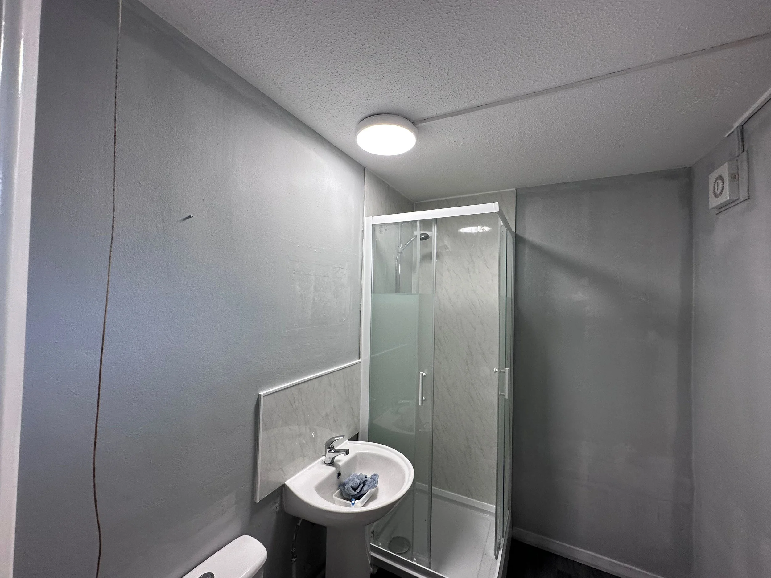 Bathroom with gray walls, a white sink with a faucet, a mirror, a towel inside the sink, shower with transparent door, and a ceiling light.