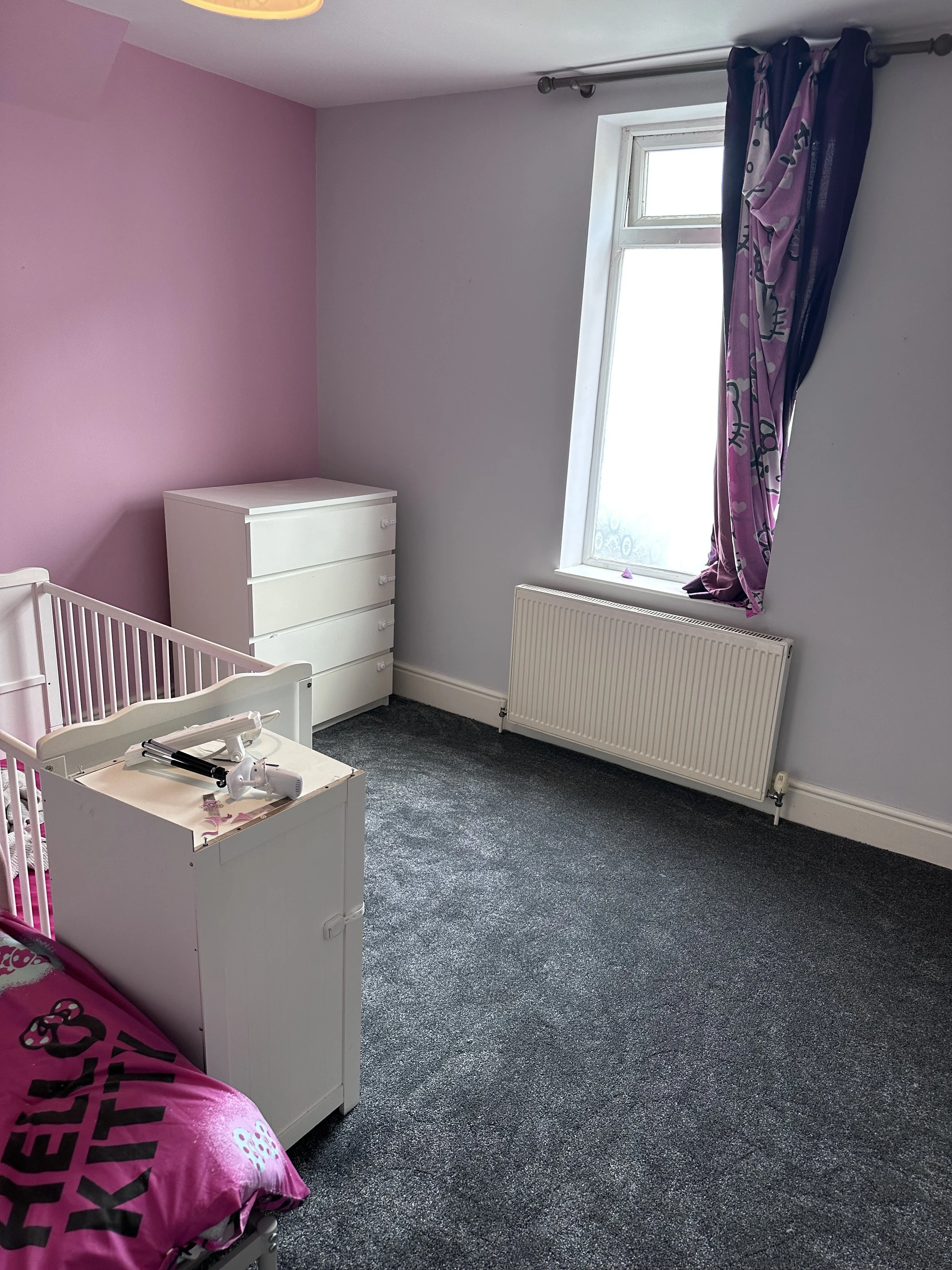 A child's bedroom with one pink wall and one light gray wall. The room has a white dresser, a pink Hello Kitty-themed pillow on the bed, a window with purple Hello Kitty curtains, and a gray carpet. There is a white radiator below the window.