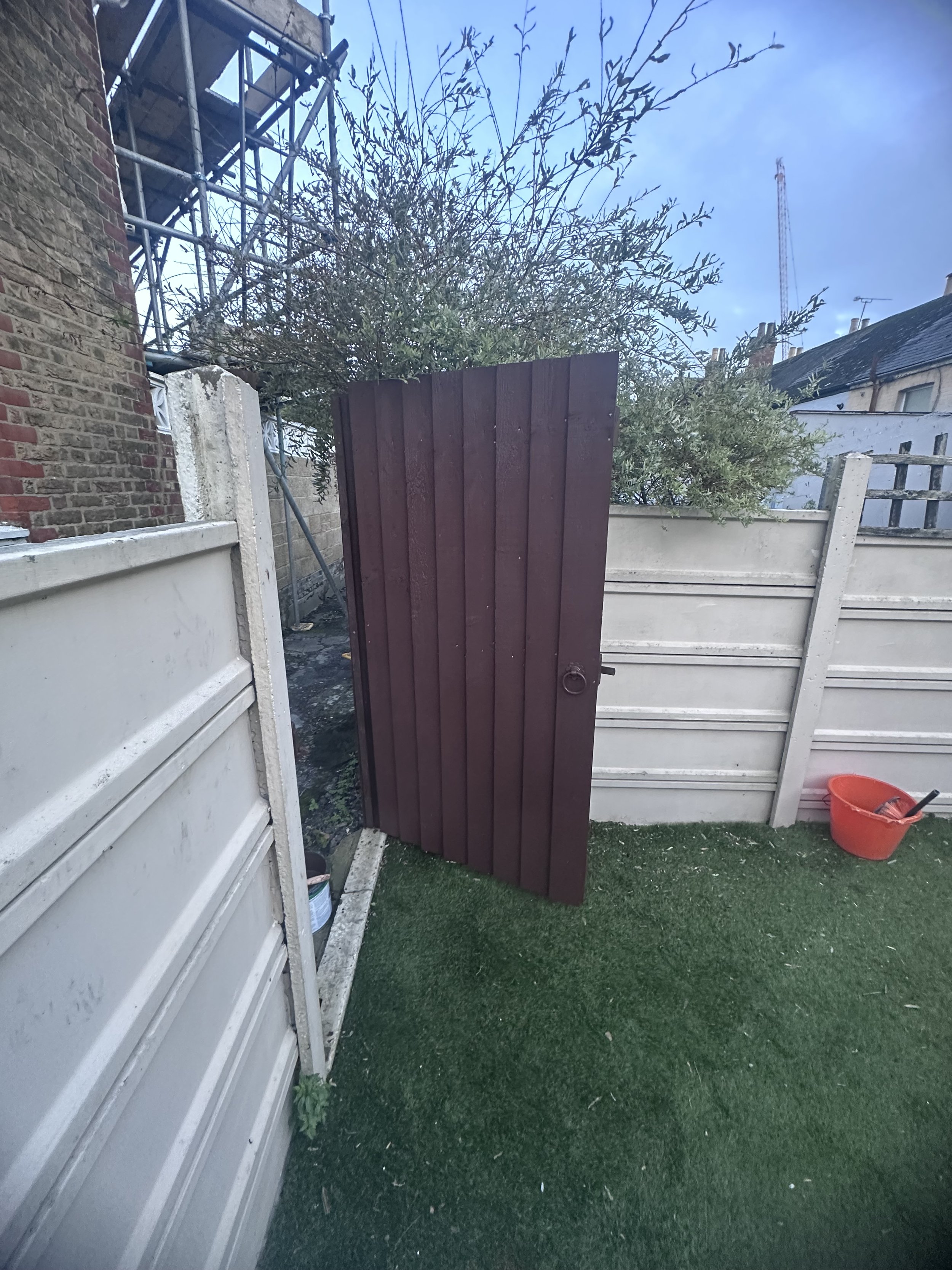 A partially assembled wooden gate leaning against a split fence in a backyard. The yard has artificial grass, and there is a red plastic bucket with tools to the right. In the background, trees and neighboring houses are visible.