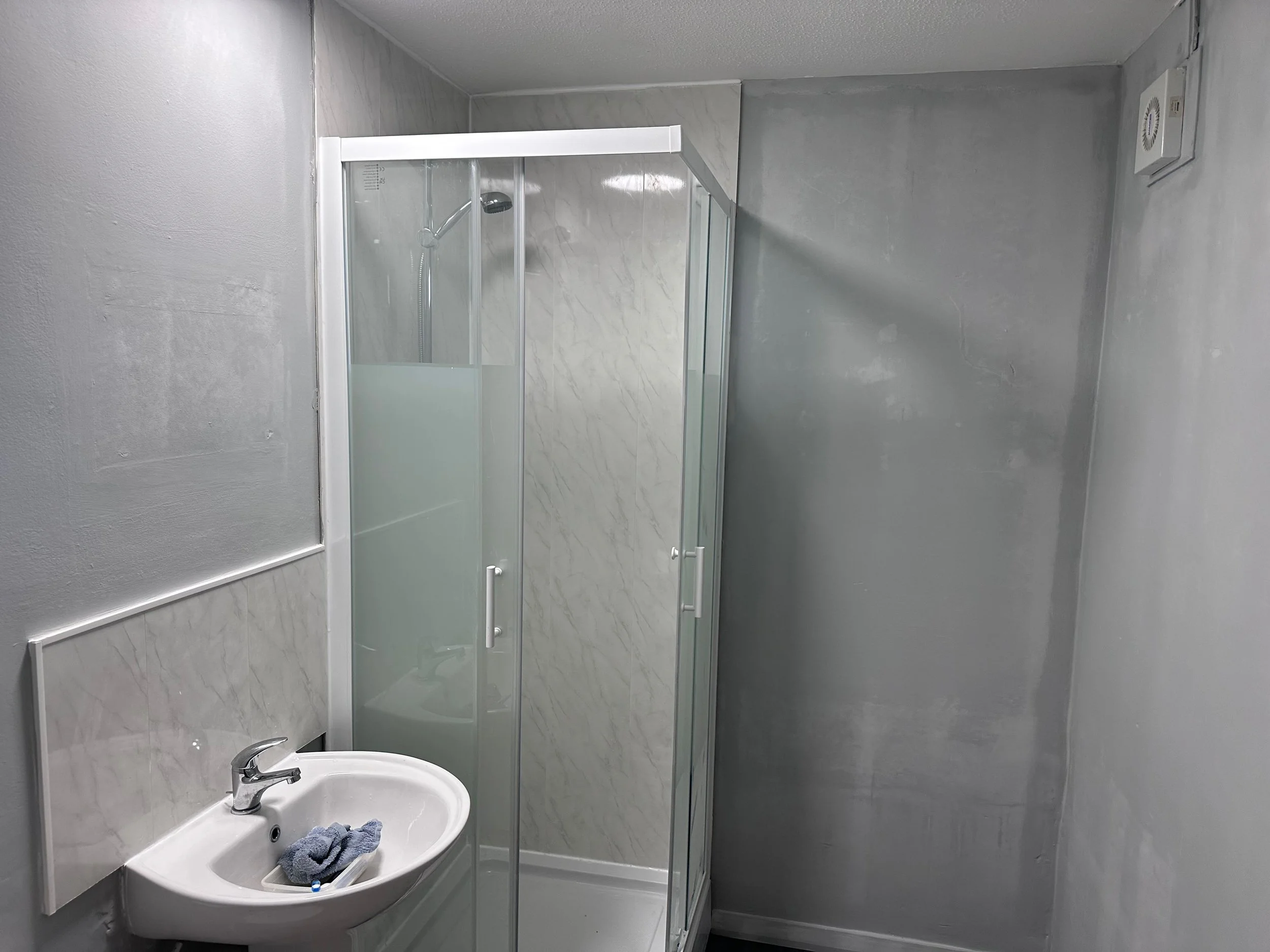 Small bathroom with gray walls, a white sink with a blue cloth on it, a mirror, a glass shower door, and an exhaust fan on the ceiling.