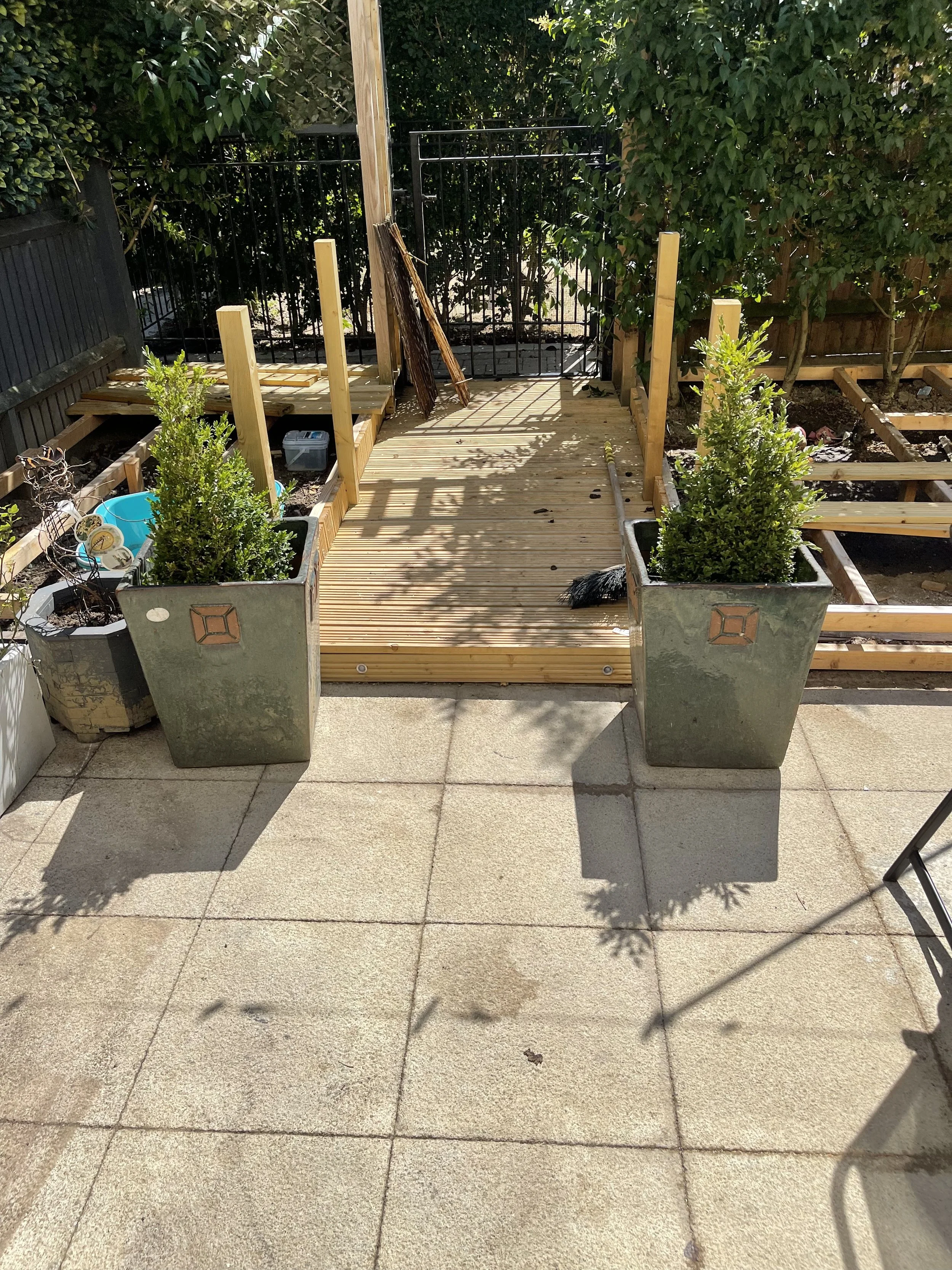 A newly built wooden deck on a concrete patio with potted plants, tools, and materials for construction. The deck is surrounded by a black metal fence and lush green trees.