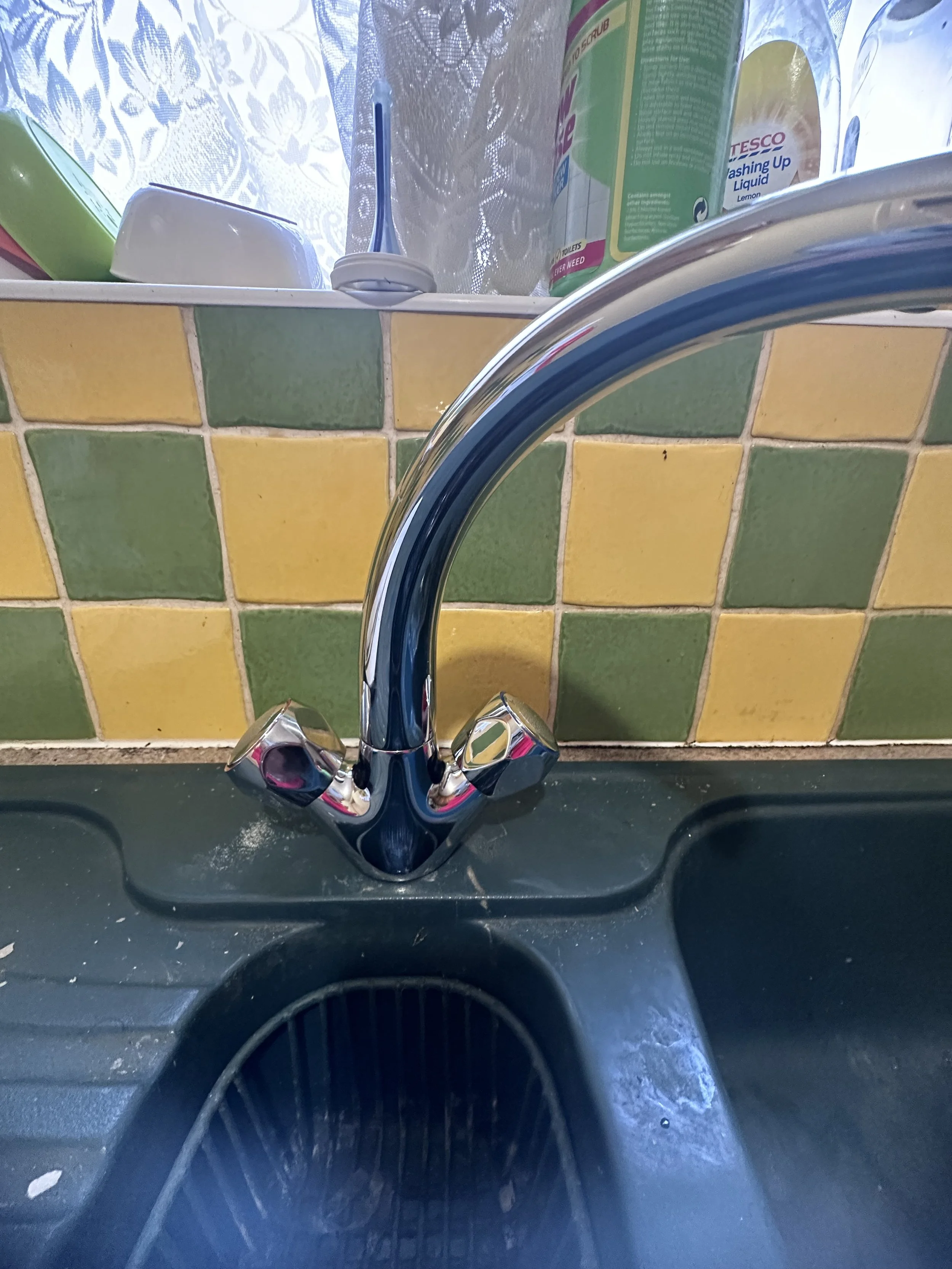 Kitchen sink with a chrome faucet, yellow and green tiled backsplash, and various cleaning products and soap in the background.