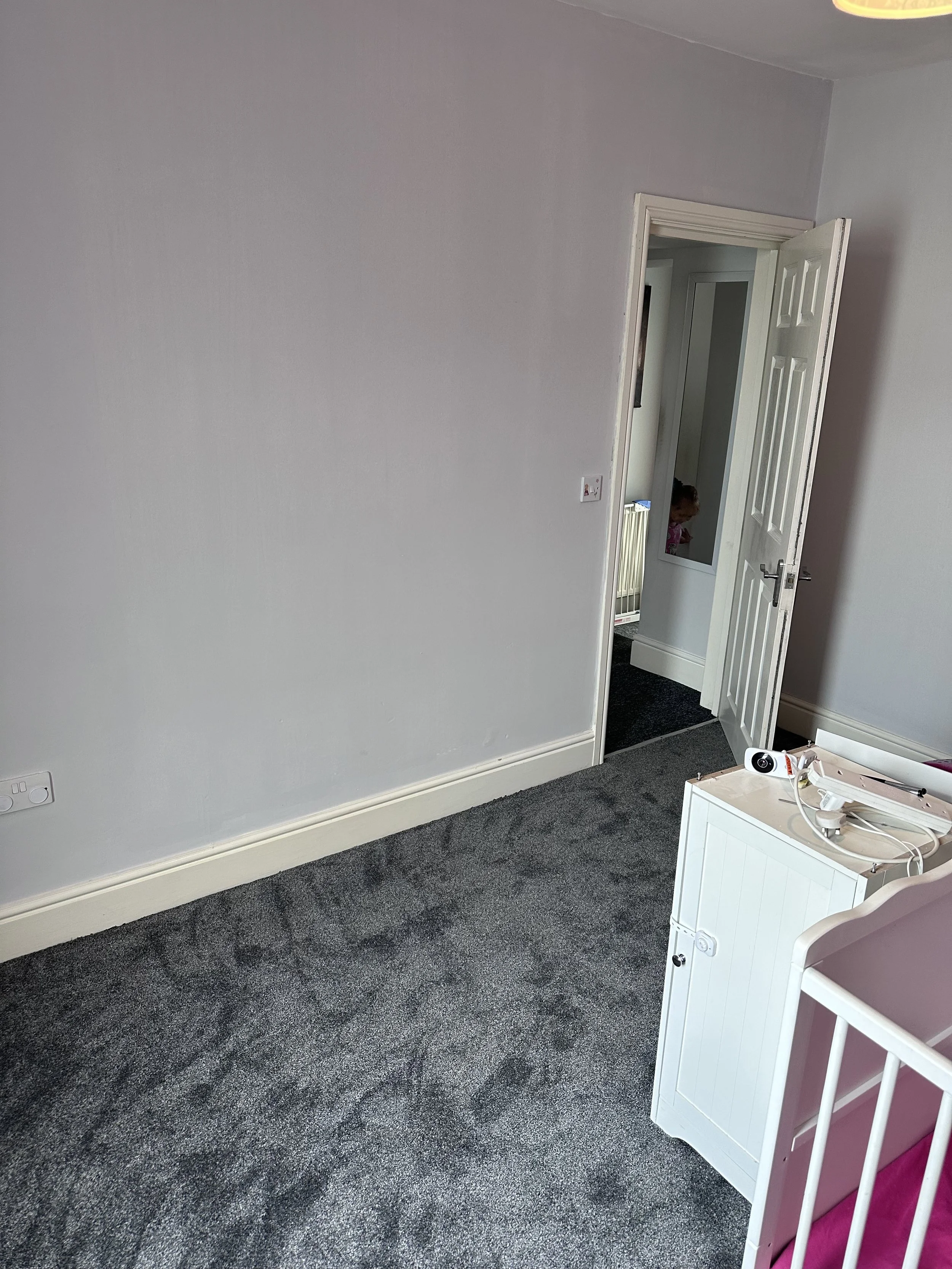 Empty room with gray carpet, white walls, open door to another room, small white furniture with a drone on top, visible child in the doorway.