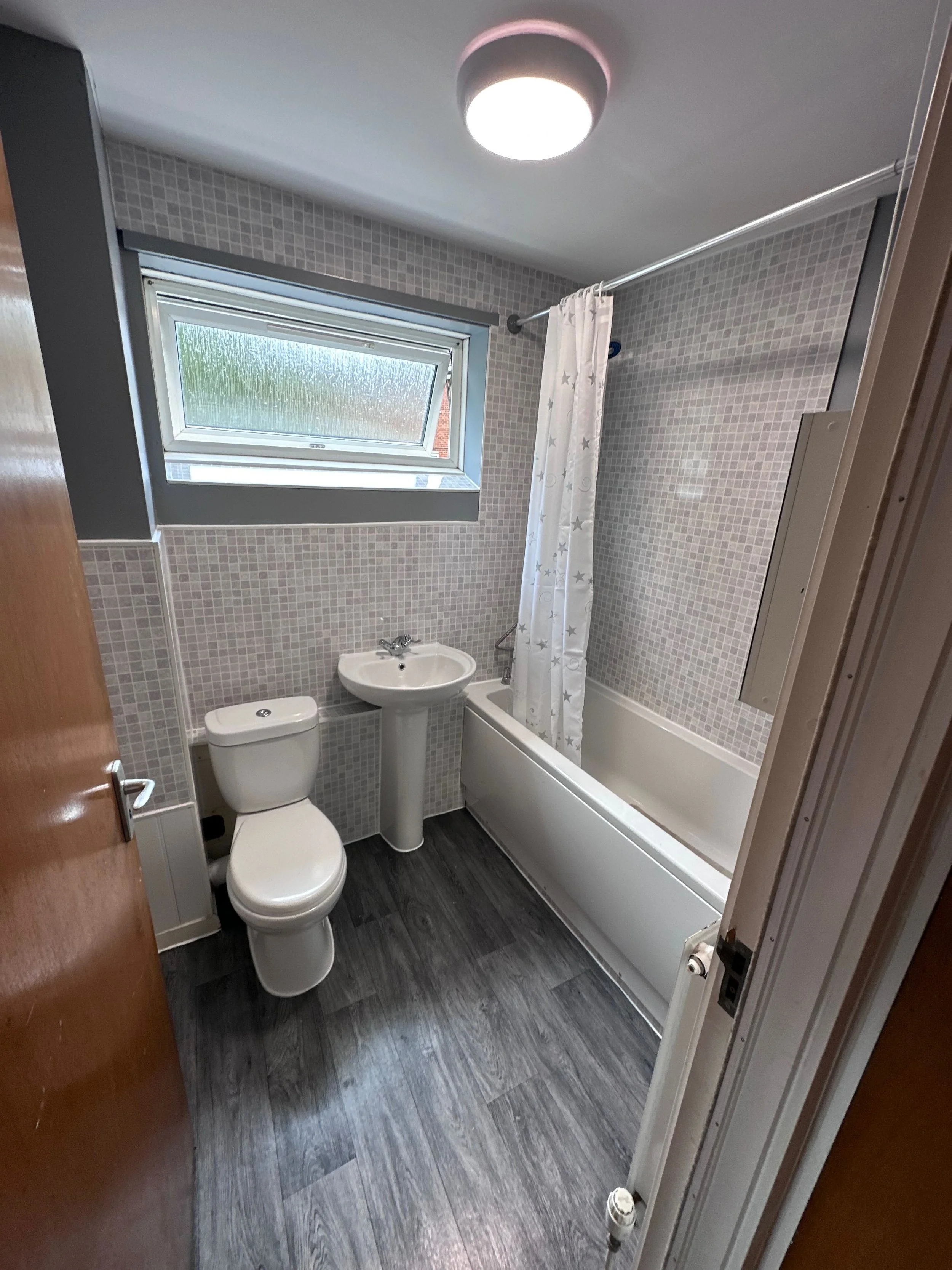 A small bathroom with a toilet, a sink, a bathtub with a shower curtain, a window with frosted glass, and a ceiling light. The floor has dark wood-look vinyl planks.