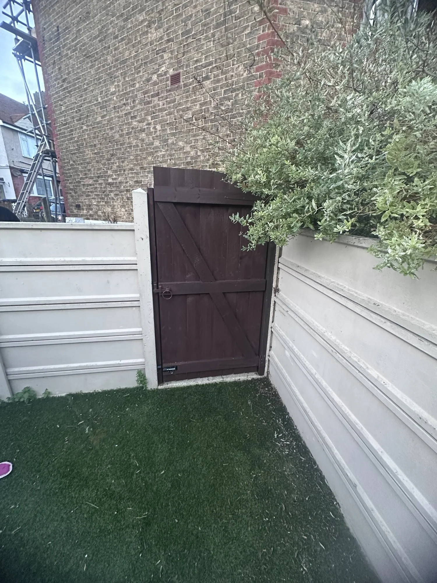 Small backyard with artificial grass, a white fence, and a brown wooden gate. There is a green bush on the right side and some debris on the artificial grass.