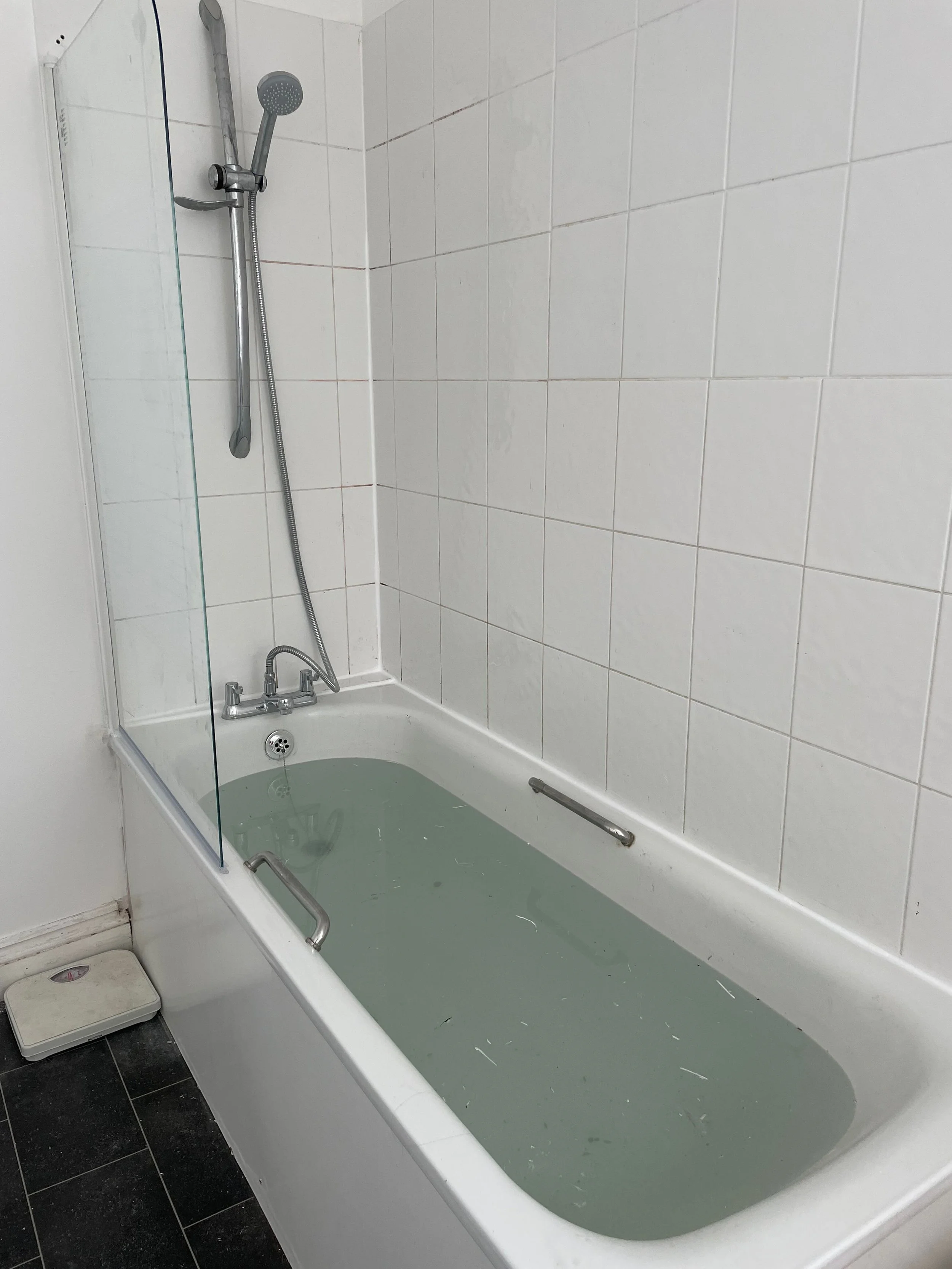 A medical bathtub filled with water, equipped with grab bars, a showerhead, and white tiled walls, in a hospital or care facility bathroom.