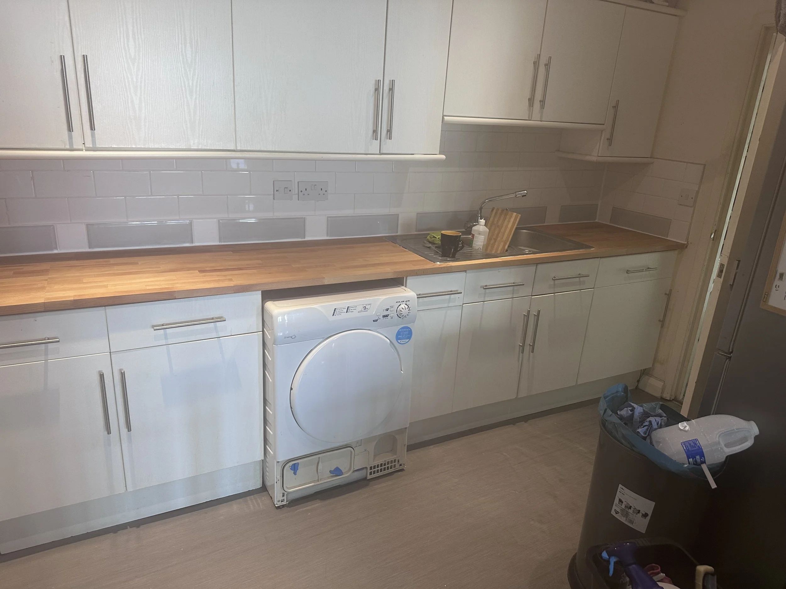 A kitchen with white cabinets, a wooden countertop, a stainless steel sink, and a washing machine. There are some items on the countertop, including a mug, a bottle of soap, and a cutting board. A trash bin with clothes inside is visible in the foreground.