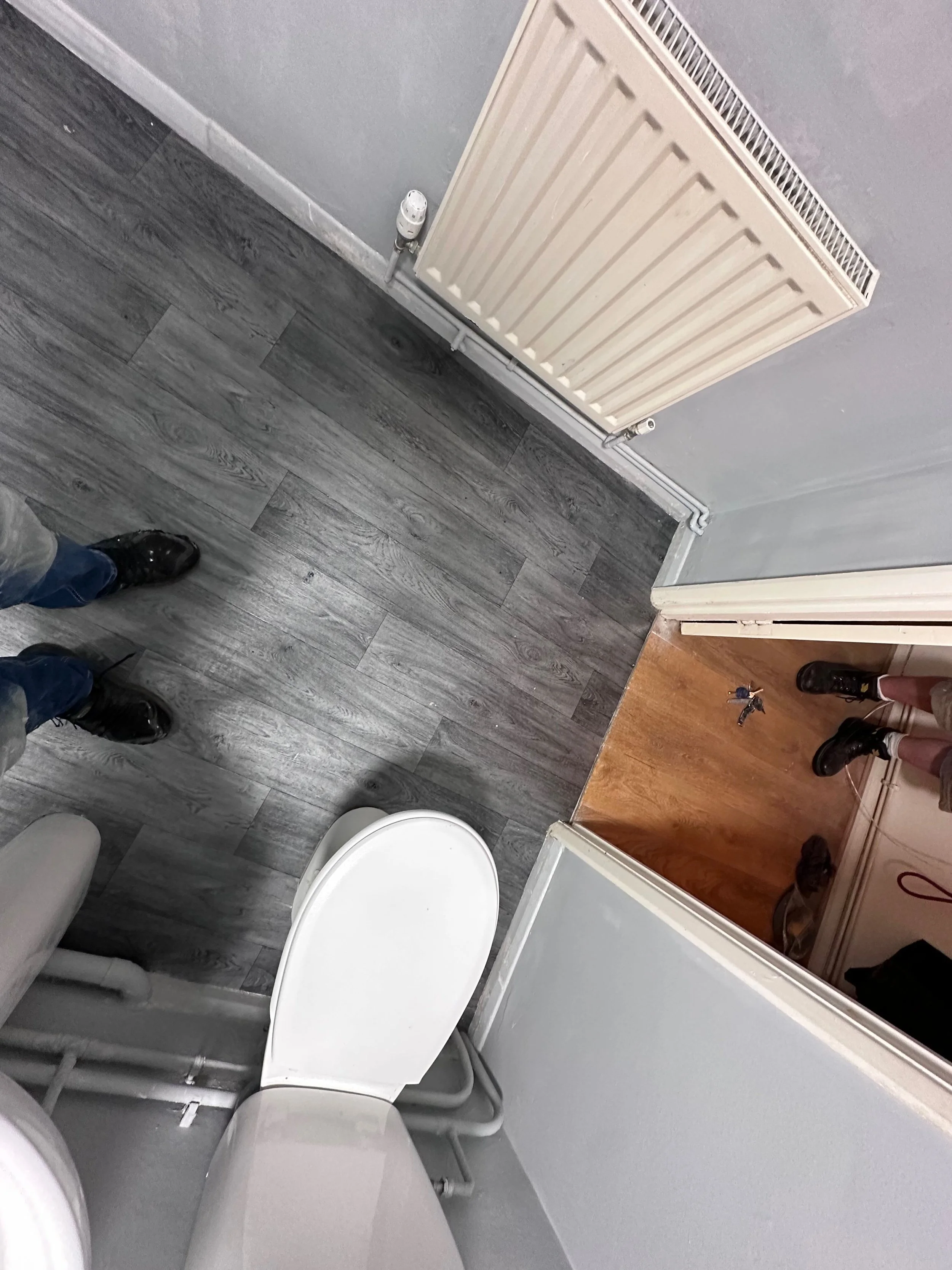 Public restroom with gray wood-look flooring, a white toilet, gray wall, radiator, and footwear of two people visible.