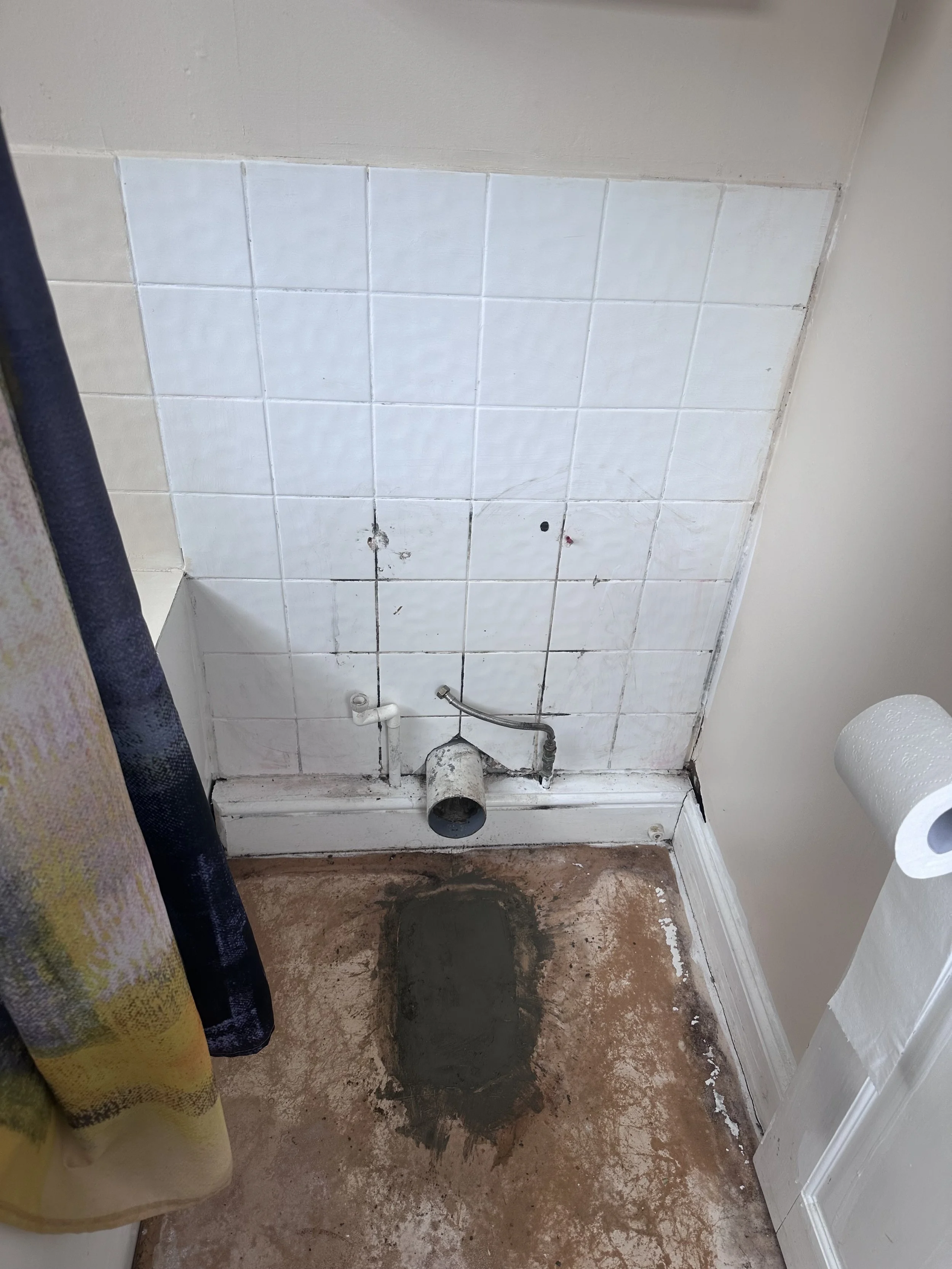 A partially renovated bathroom shows white tiles on the wall, exposed pipes, and a brown, dusty floor. There's a shower curtain on the left, and the room appears to be under construction or repair.