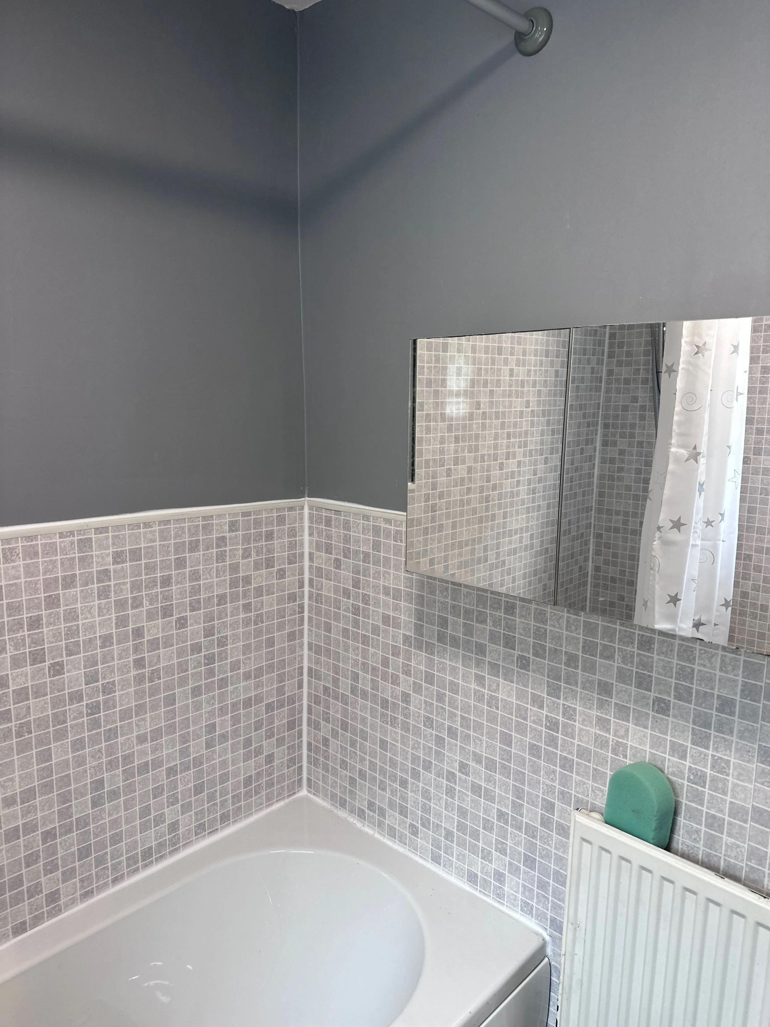 A bathroom corner with a bathtub, gray tiled walls, a mirror, a shower curtain with stars, a green soap dish, and a white radiator.