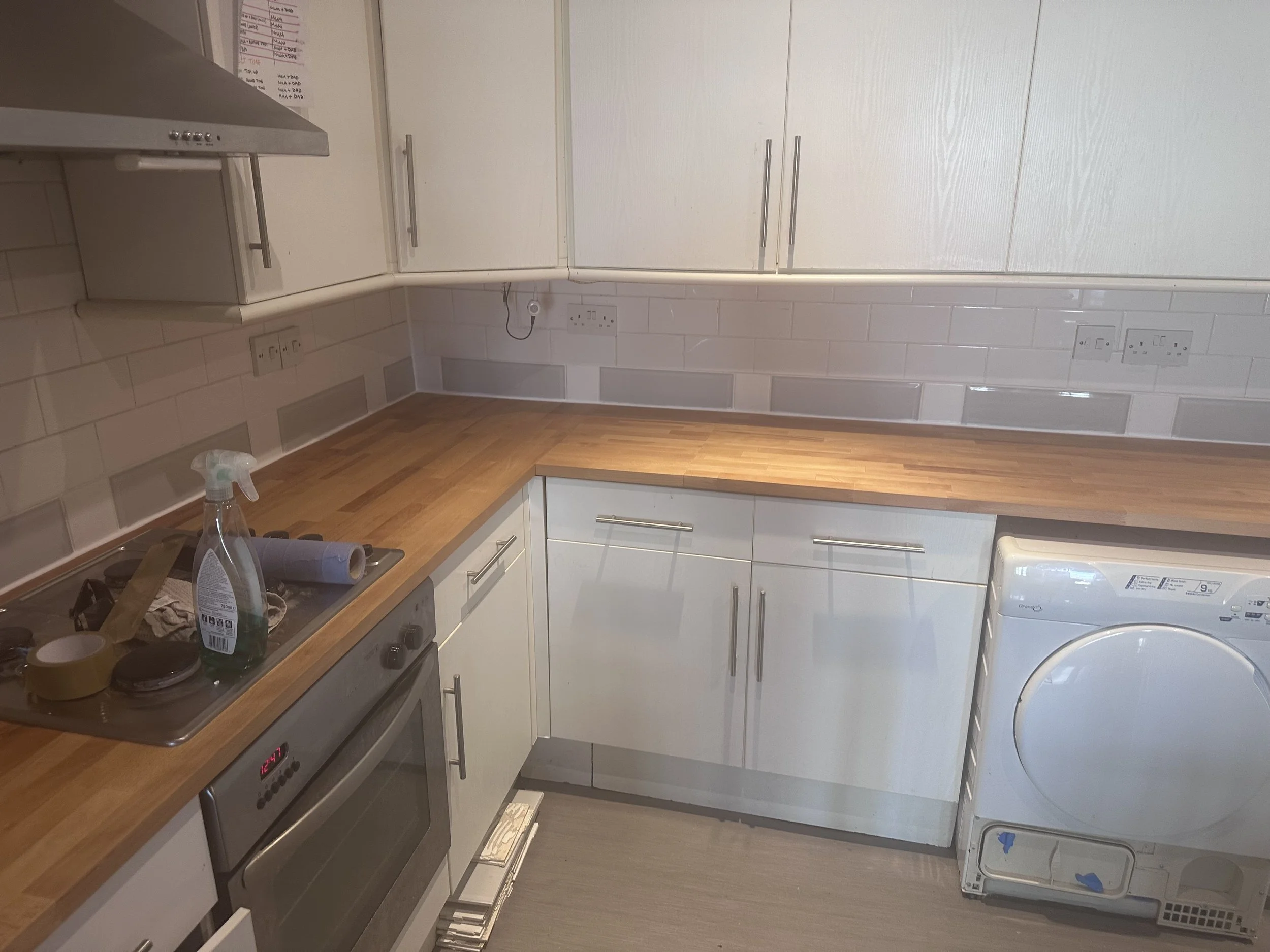 A small kitchen with white cabinets, a wooden countertop, a gray stove, and a white washing machine. There are electrical outlets and a tiled backsplash on the wall.