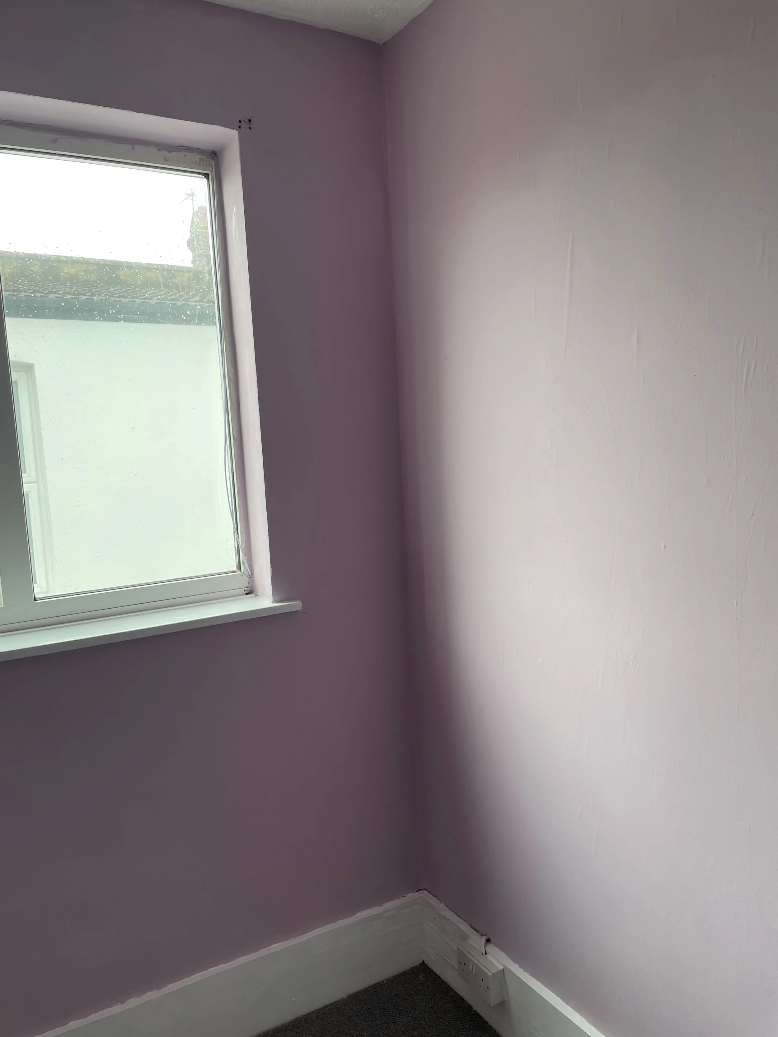 Interior corner of a room with lavender painted walls, a window letting in natural light, white window sill, white baseboard, and a dark carpeted floor. An electrical outlet is visible near the floor on the right wall.