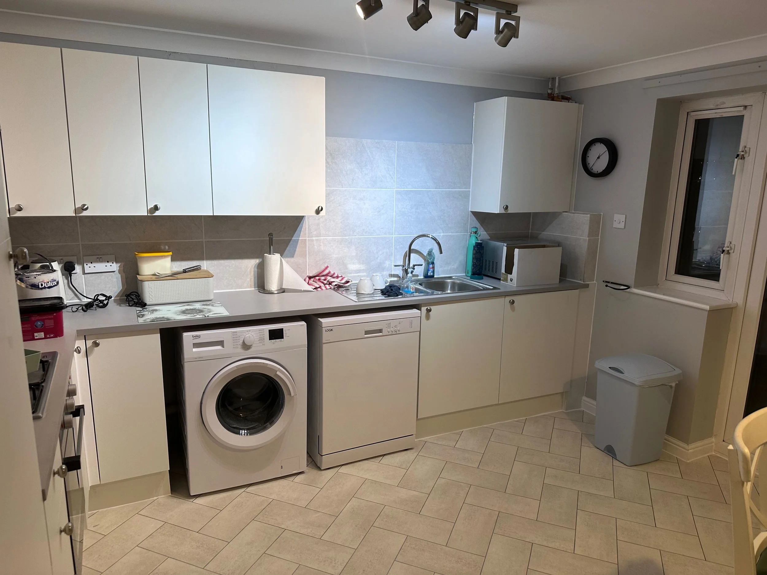 A cozy kitchen with white cabinets, a countertop, and various small appliances, including a microwave and a washing machine. There are cups, bottles, and kitchen towels on the counter. The room has tiled floors, a window, and a wall clock.