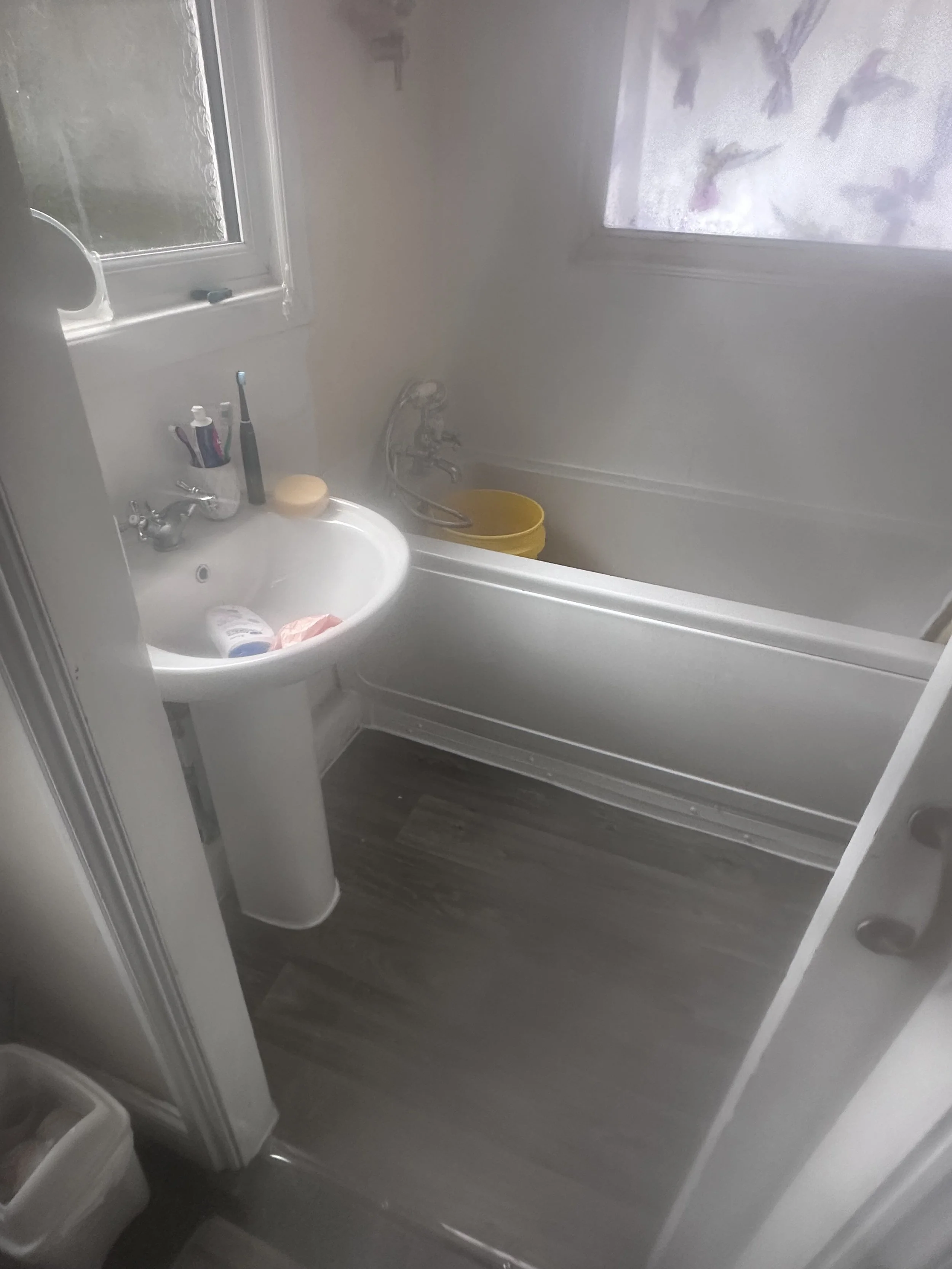 A small bathroom with a white pedestal sink, toothbrushes, a tube, a soap dish with soap, a yellow bucket in the bathtub, and a window with frosted glass.