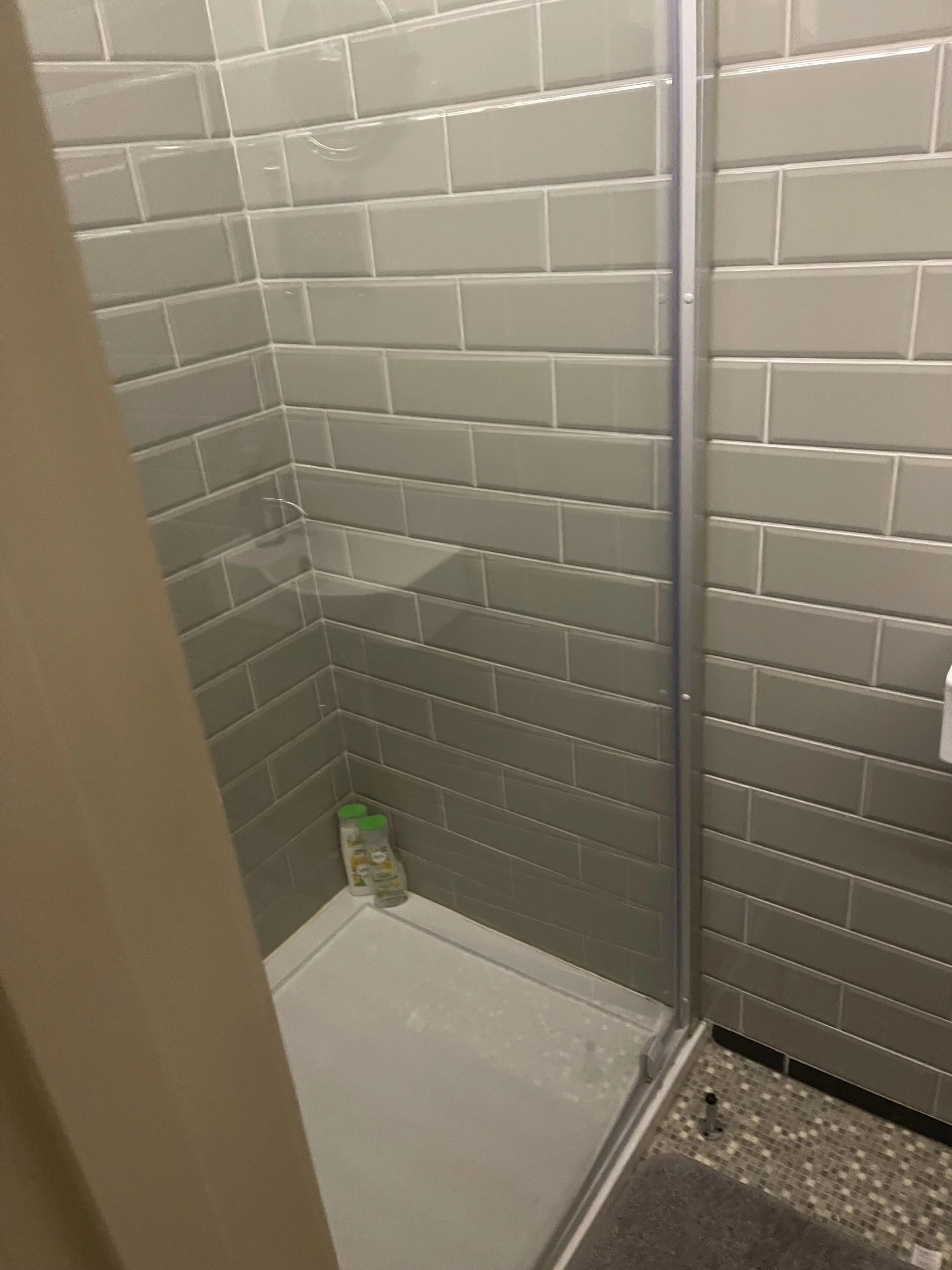Inside a shower with gray tiles, a bottle of shampoo or body wash is placed on the shower floor near the corner.