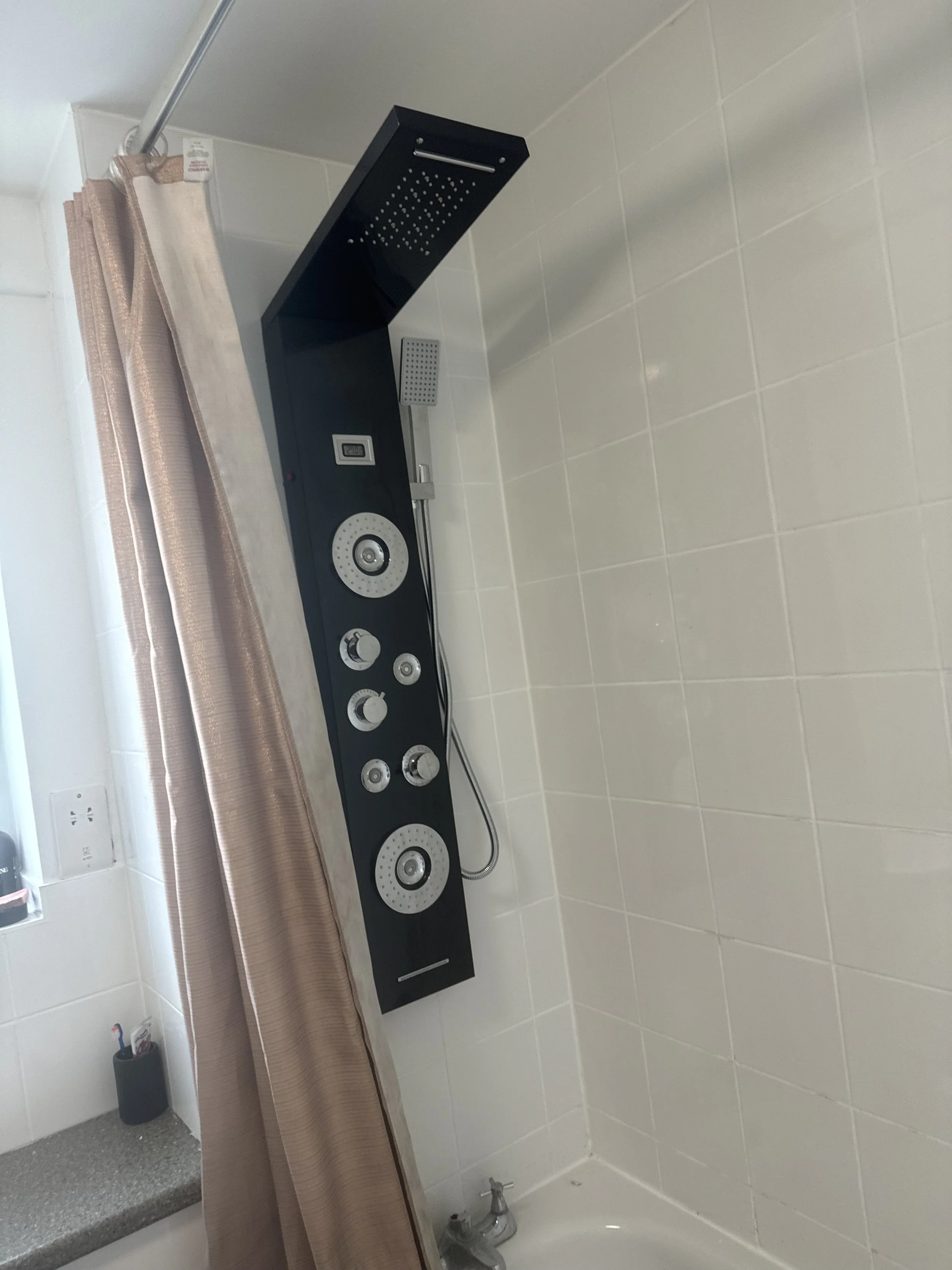 Bathroom with tiled wall, black shower panel with multiple knobs, a small grey shower head, a pink shower curtain, and a gray countertop with a black container and an electrical outlet.