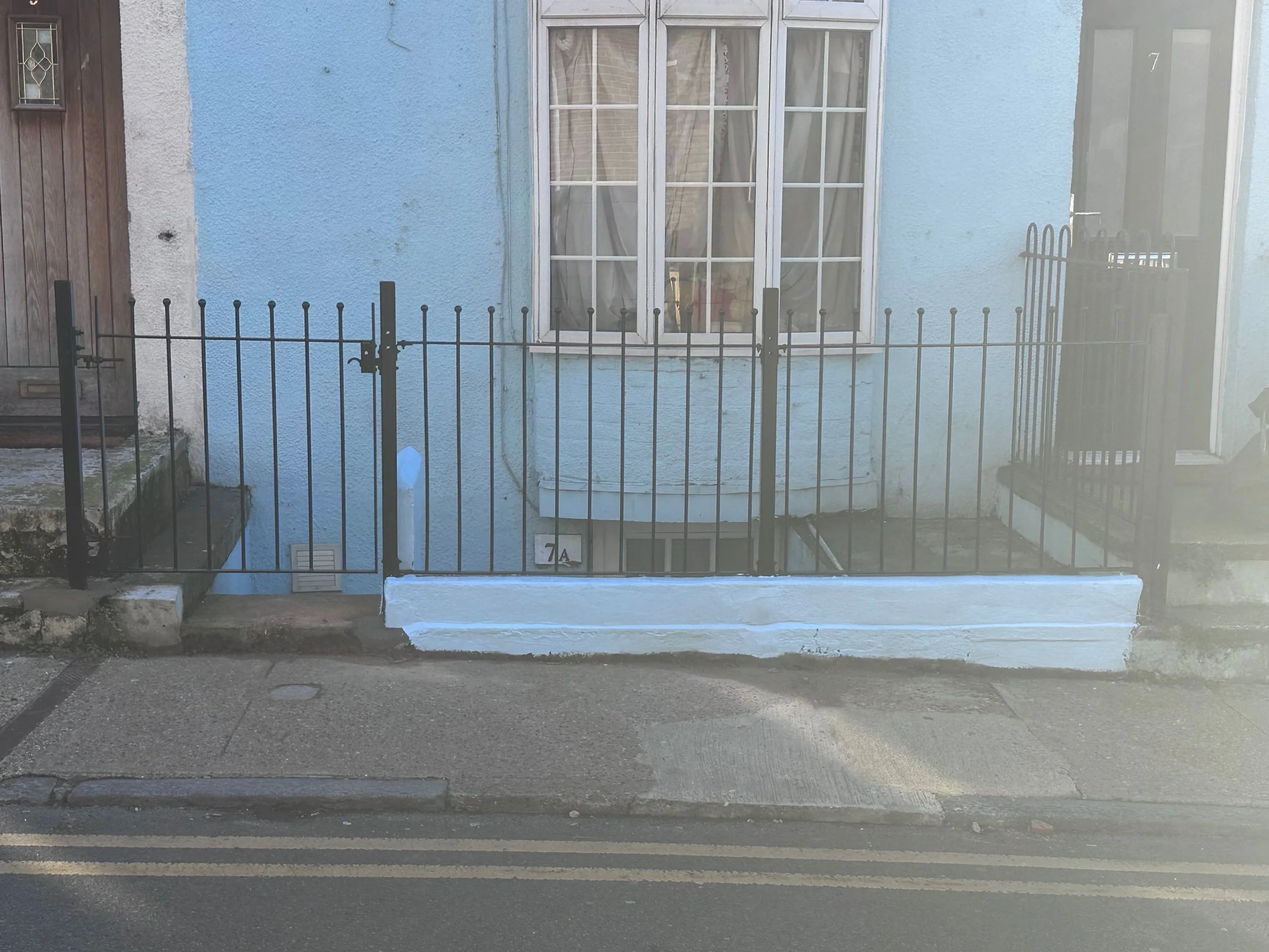 Photo of a house with a blue stucco wall, a white-framed bay window with curtains, a small front yard with a black metal fence, concrete steps, and a sidewalk.