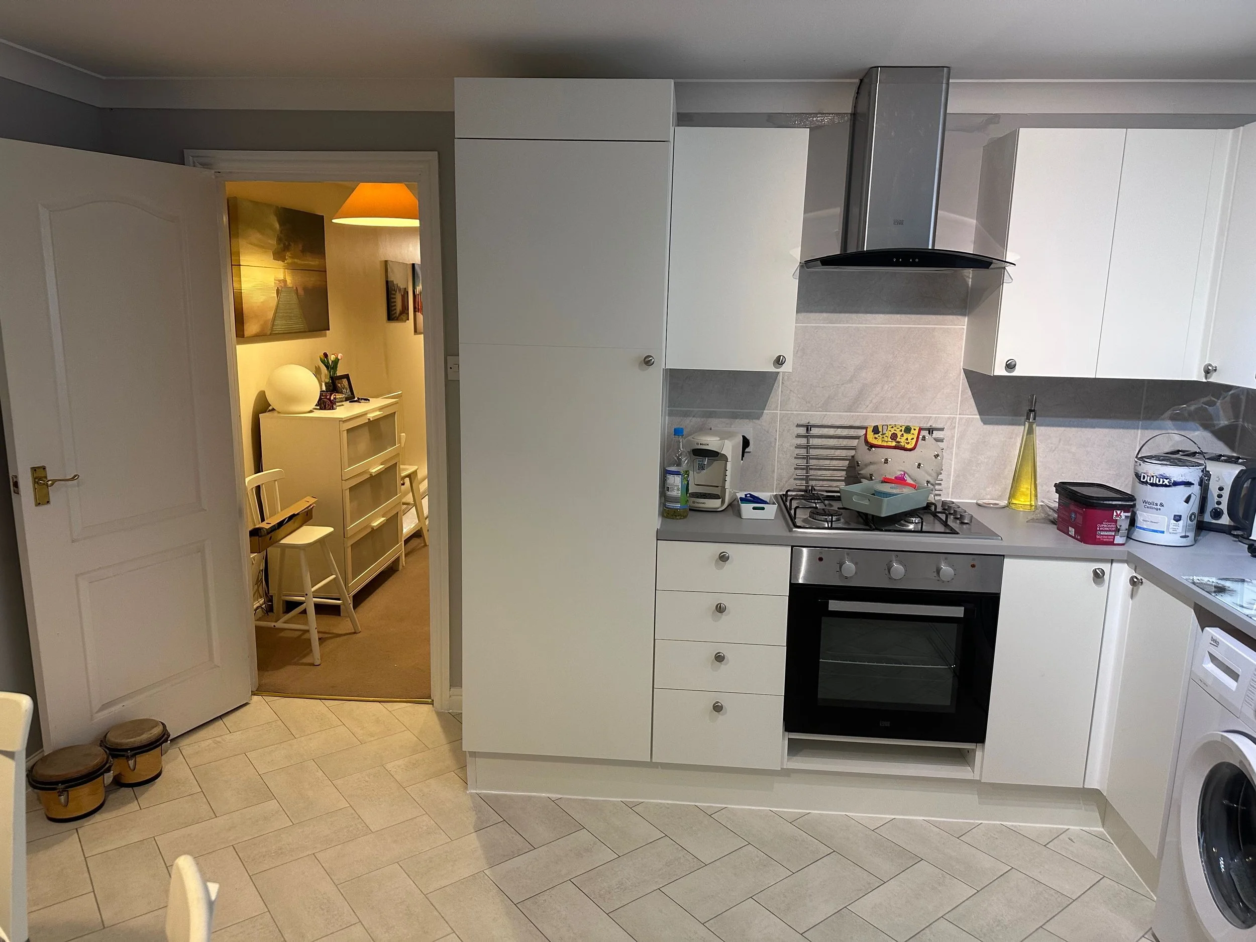 A kitchen with white cabinets, a stainless steel range hood, a stove, and various kitchen items on the countertop, with a doorway leading to a dining area visible in the background.