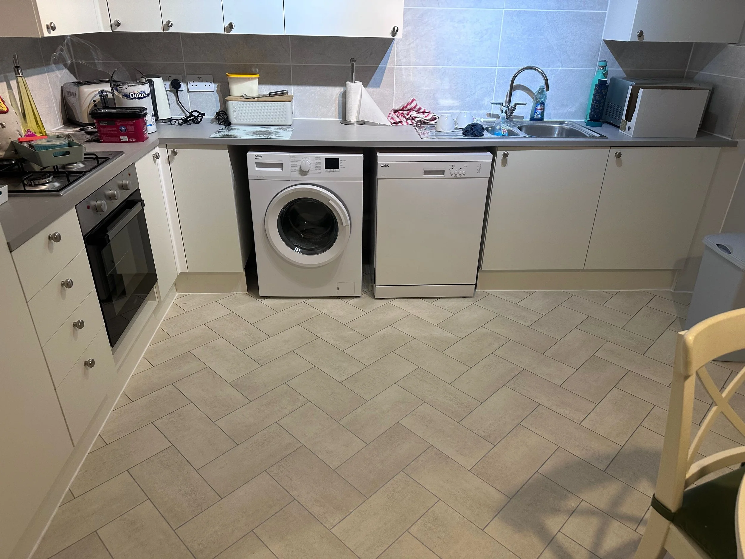 A kitchen with white cabinets, a countertop with various appliances and items, a washing machine, a microwave, a sink with cleaning supplies, and a chair partially visible in the foreground.