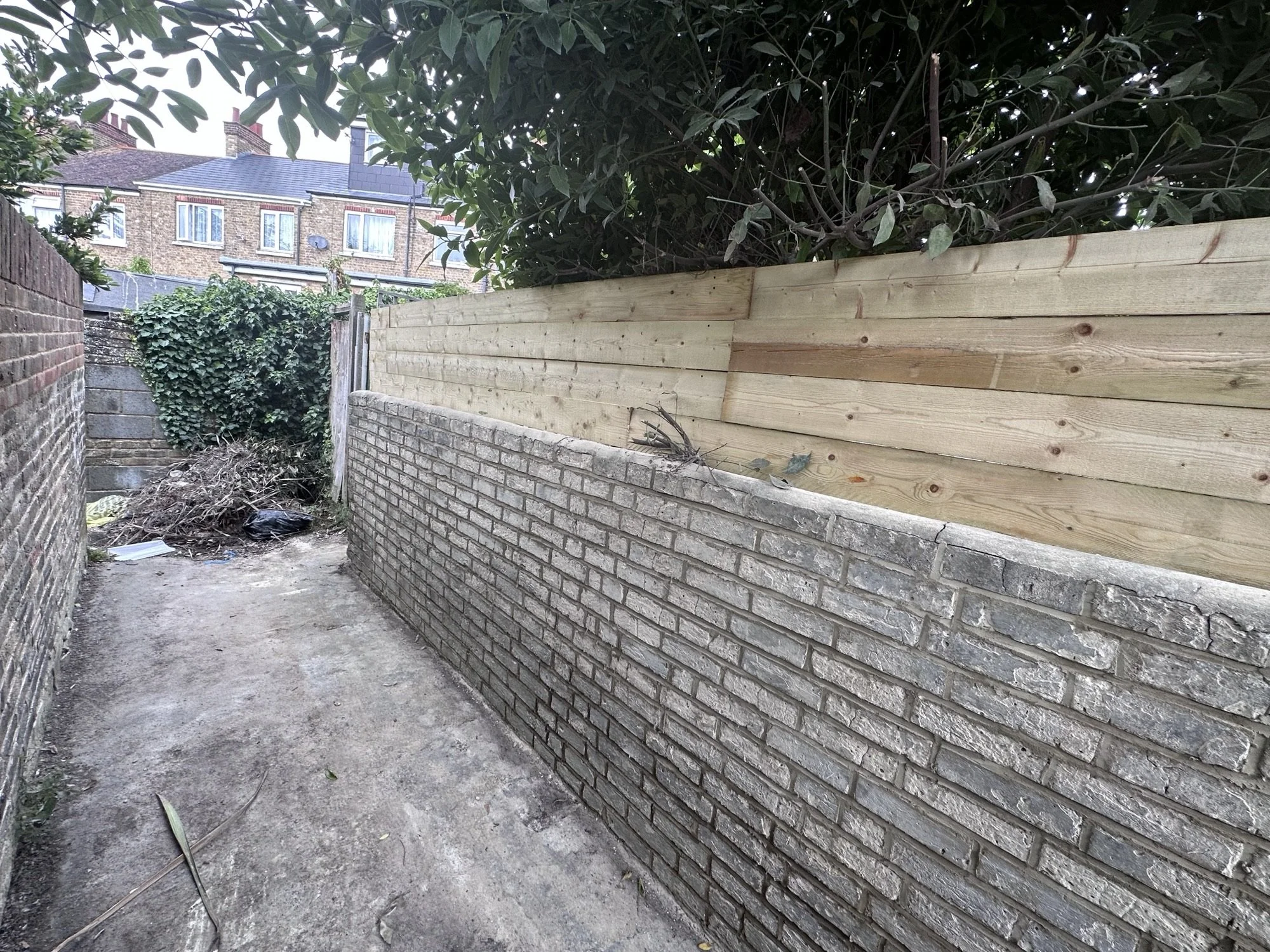 An alleyway with brick walls on both sides, with a newly built wooden fence on the right side. There is some debris and leaves on the ground, and a tree with dark green leaves overhangs the area.
