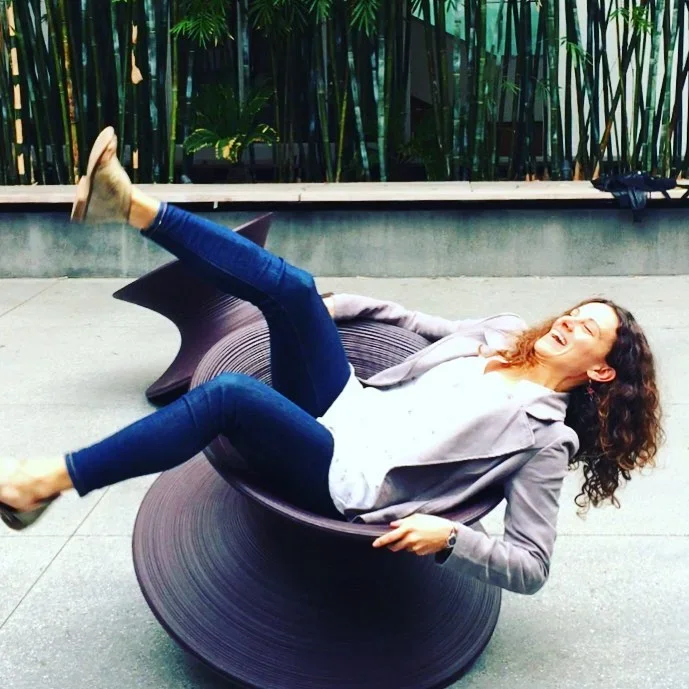 A woman with curly hair laughing while lying on a modern, curved, black outdoor chair on a concrete surface with green bamboo plants in the background.