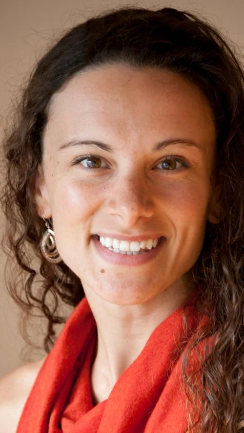 Close-up of a woman with curly brown hair, wearing earrings and a red collar smiling at the camera.