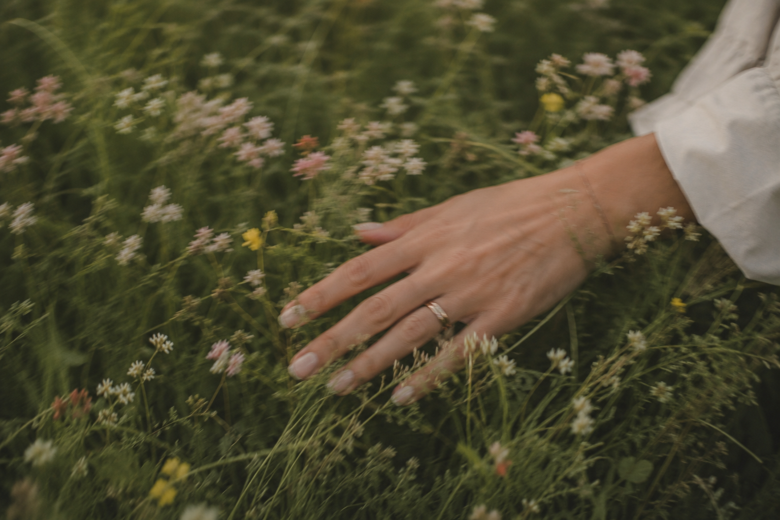 A person's hand with a wedding ring resting on wildflowers and grasses in a field.