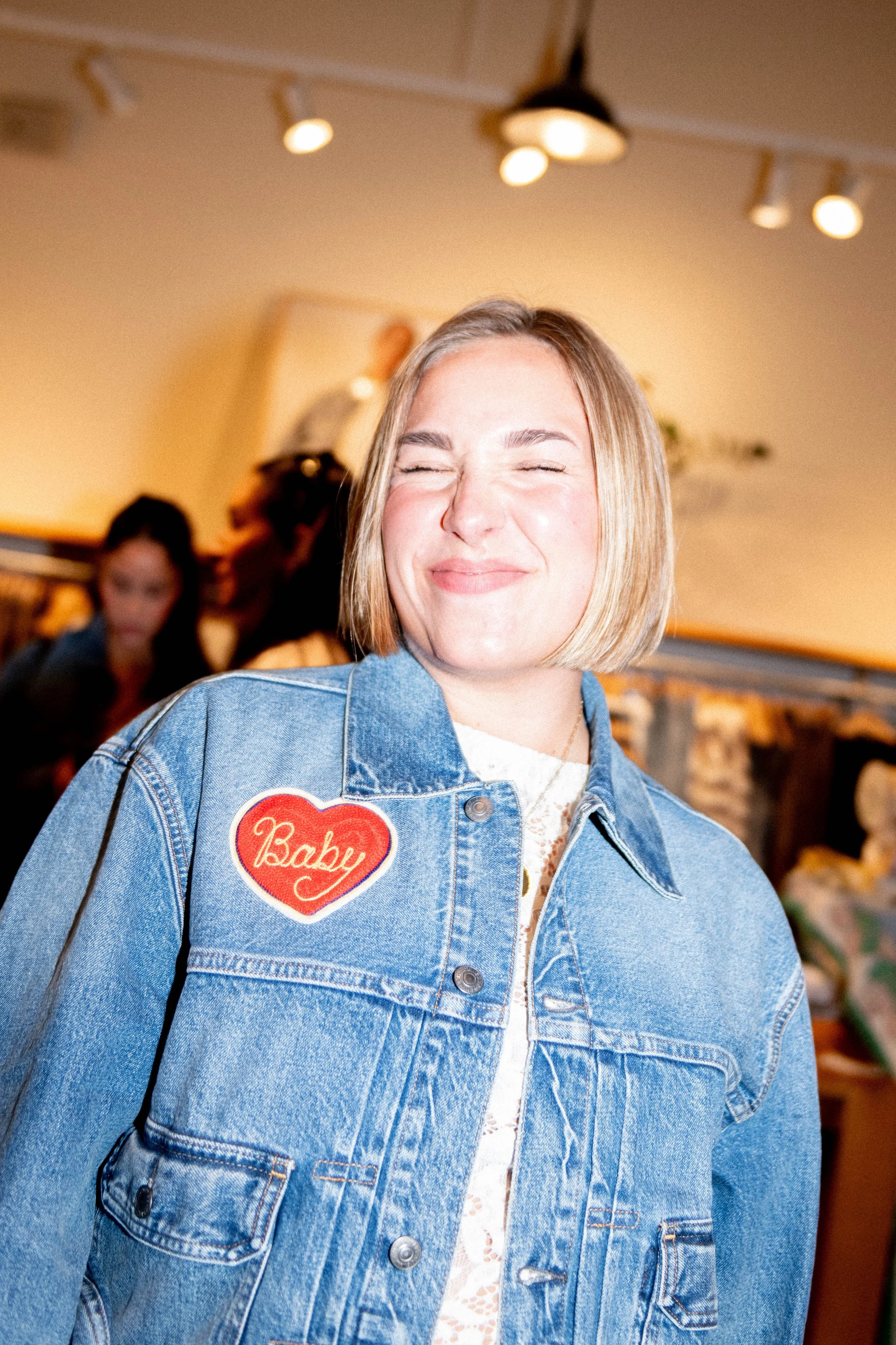 Young woman with blonde hair smiling, wearing a denim jacket with a heart-shaped patch that says 'Baby'.