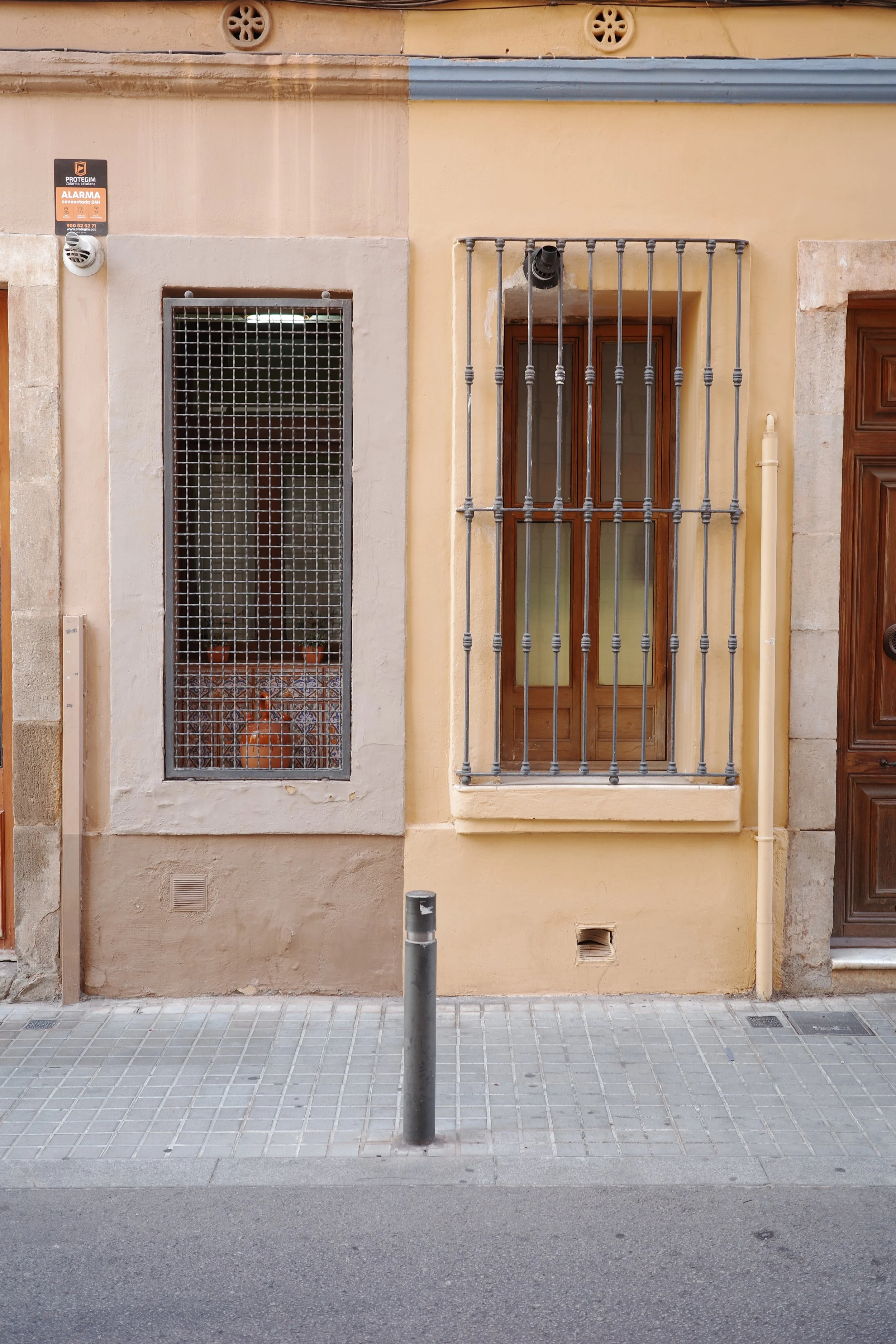 A beige building façade with two windows and one door. The left window has a metal grid, and the right window has metal bars. There are small round vents near the top of the building. A gray pipe runs vertically beside the right window, and a short b
