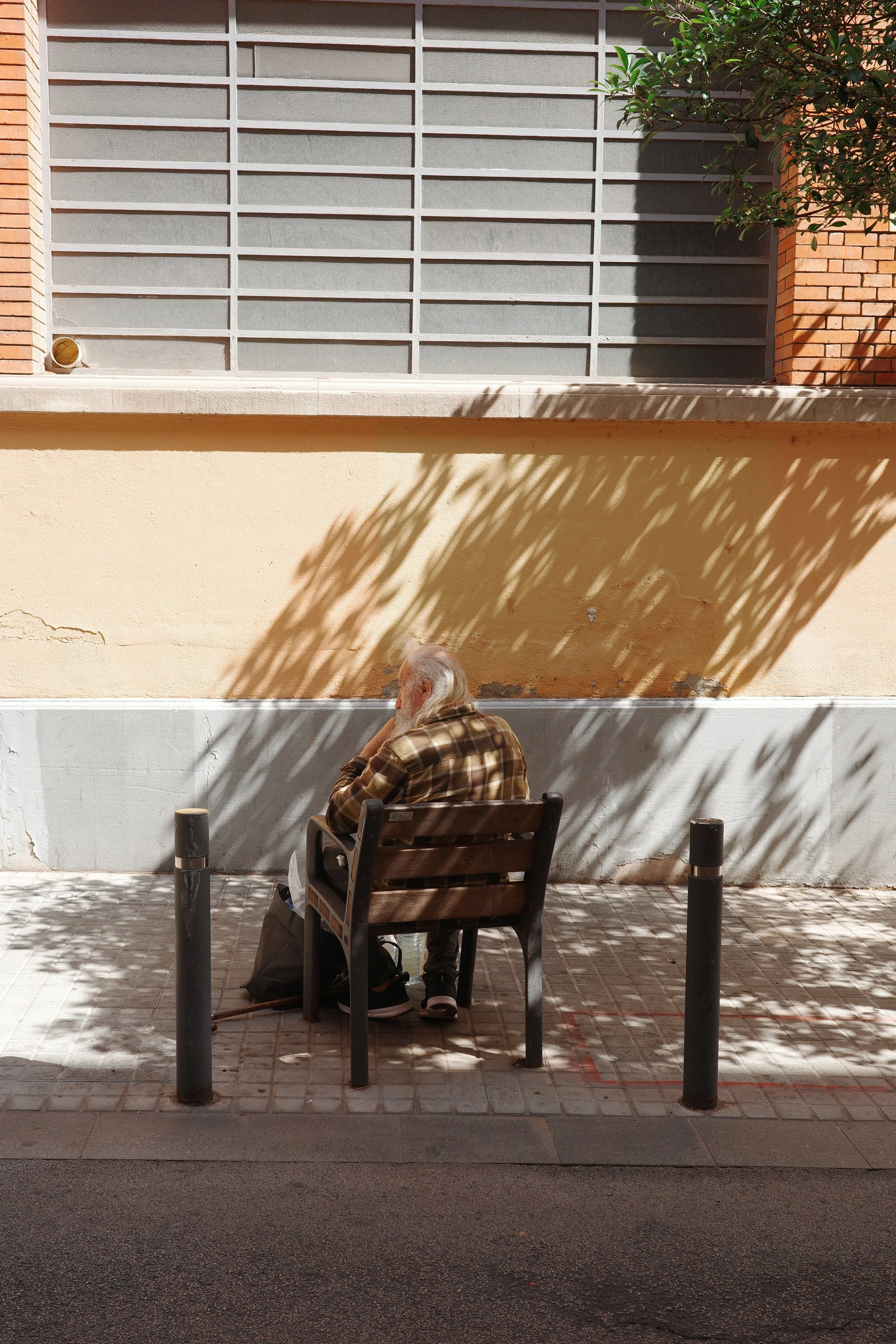 An elderly man with a walking cane sitting on a wooden bench in front of a beige wall, with shadows of leaves overhead and a building with a metal window frame nearby.