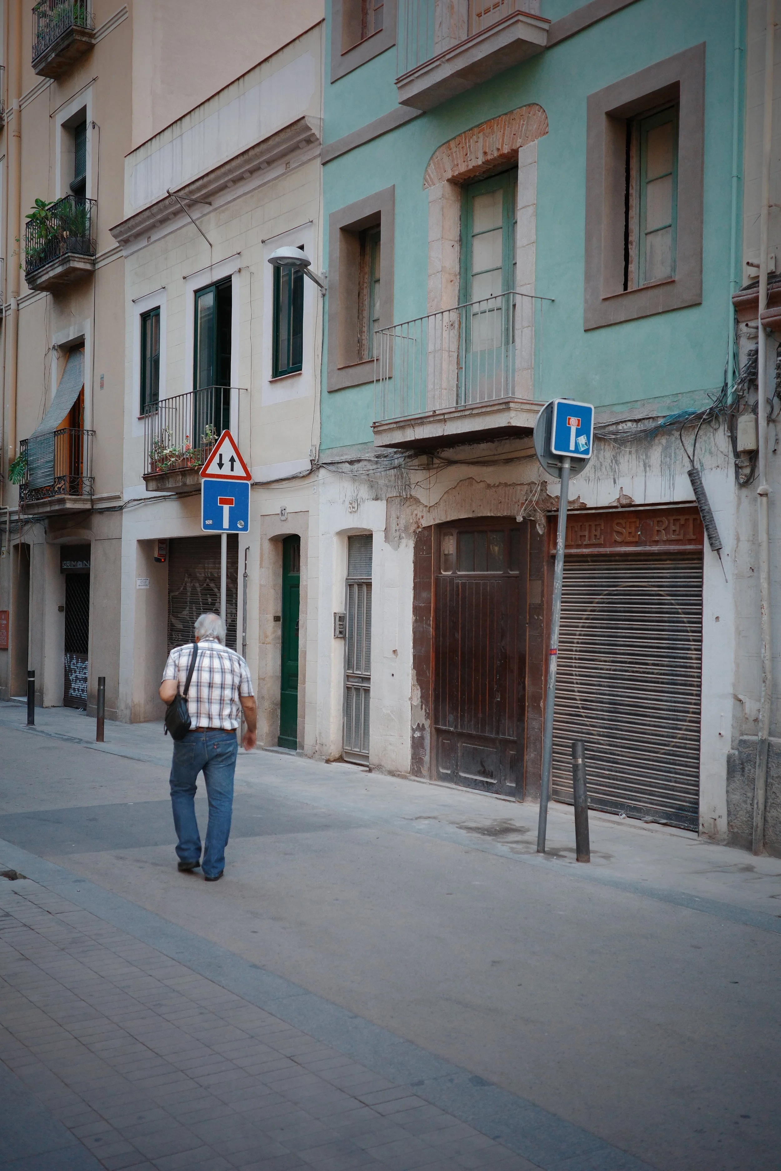 A man walking down a city street with multi-story buildings on either side. The buildings have balconies and awnings. There are signs on the street, one indicating a dead end, and another showing a one-way street. The buildings are worn, and some sto