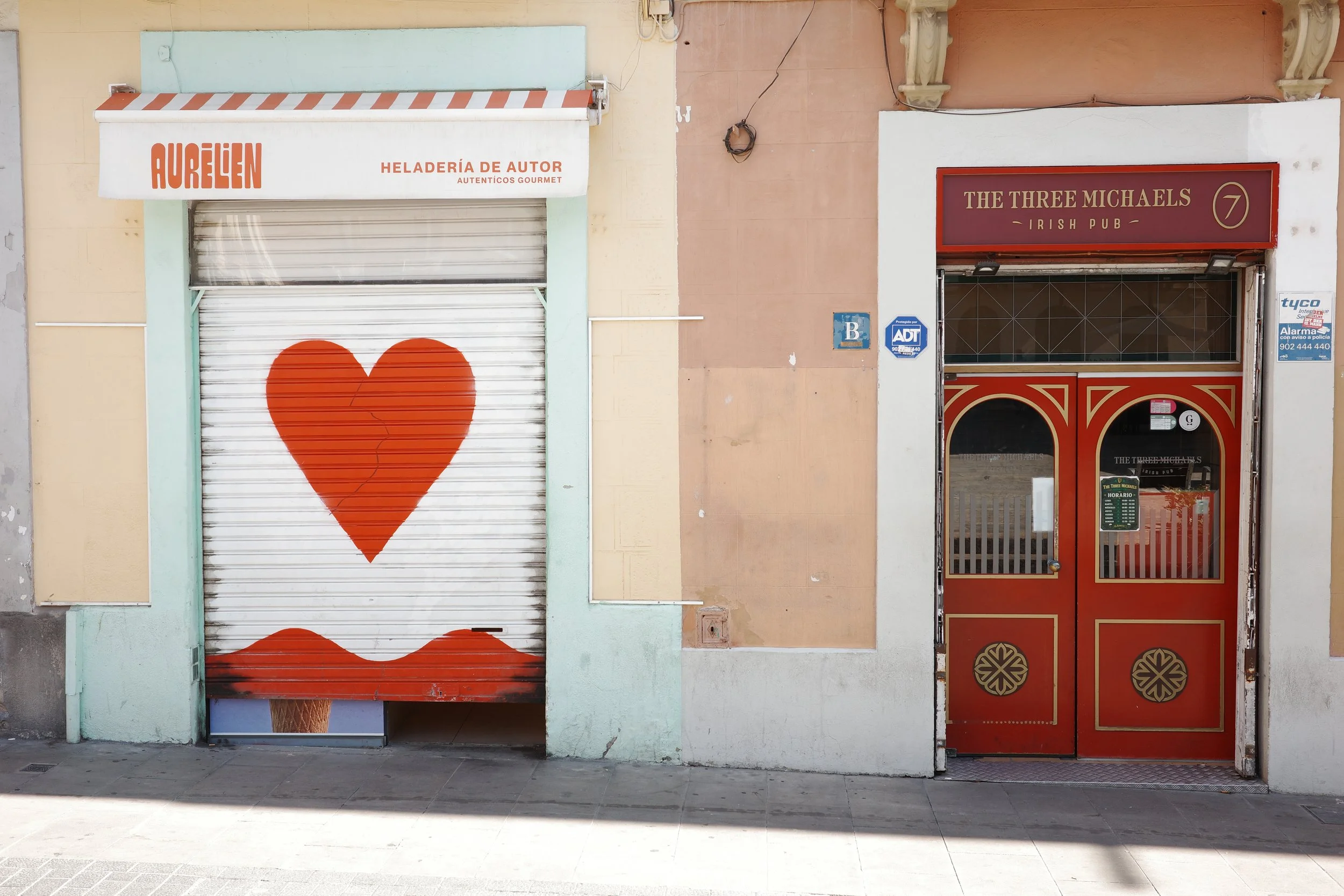 Closed storefront with a white metal shutter featuring a red heart with a crack in it, next to a door with a red sign for an Irish pub named 'The Three Michaels'.