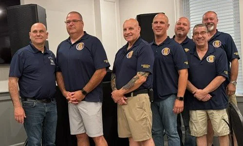 Six men standing indoors, wearing navy blue polo shirts with a logo, in front of audio equipment and white walls.