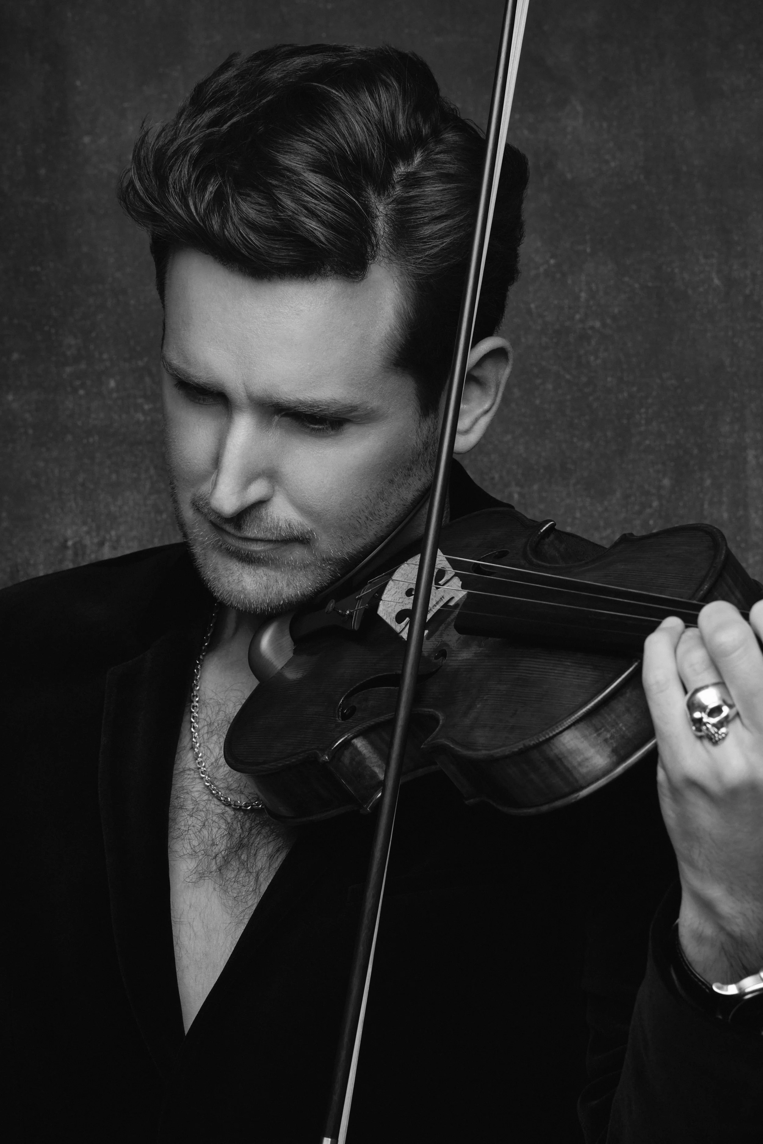 A man with dark, wavy hair plays a violin. The image is in black and white, and he is wearing a dark jacket with a chain necklace. His eyes are closed or looking down as he concentrates on the music.